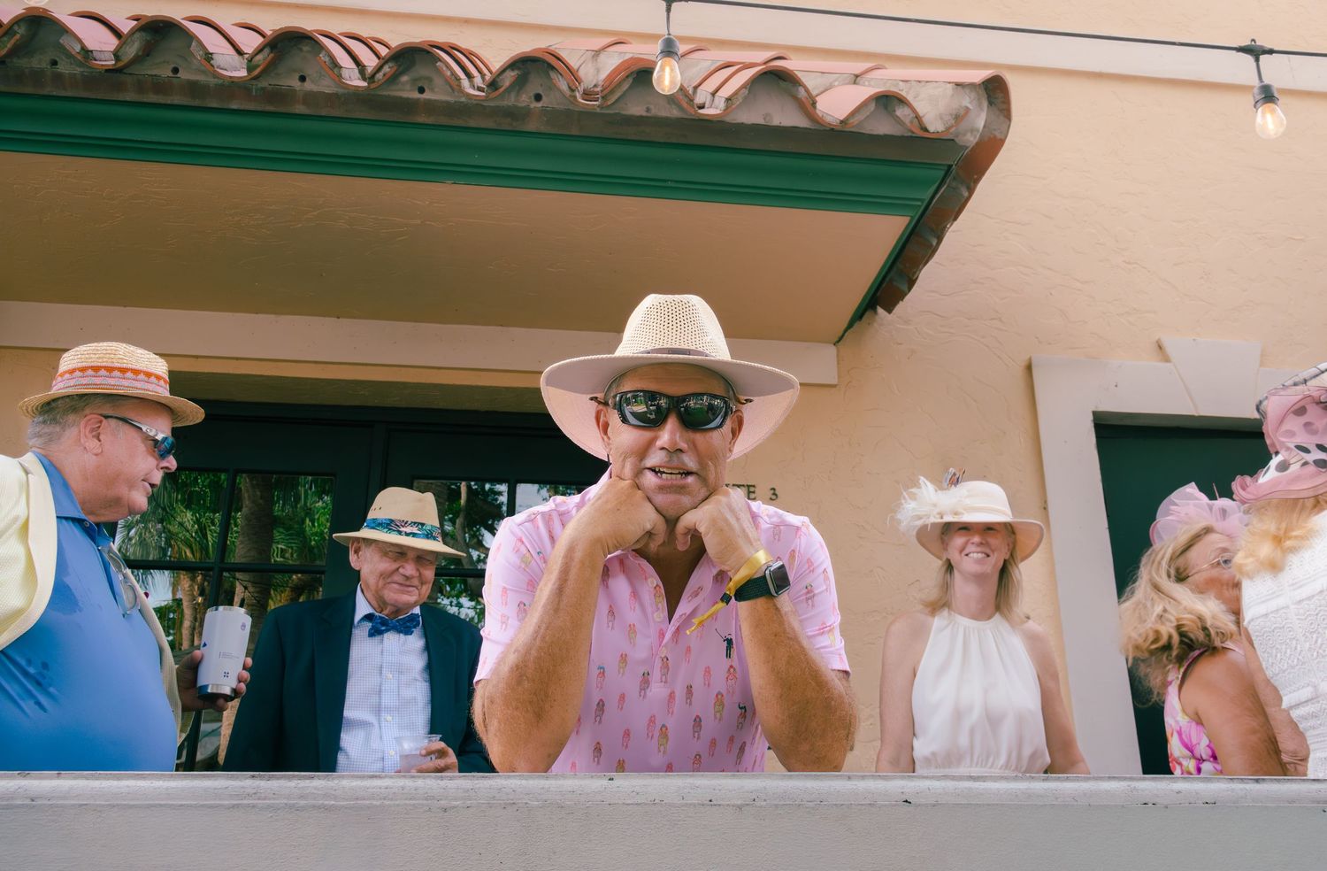 Man in pink shirt and Panama hat watching Kentucky Derby festivities from an outdoor balcony.