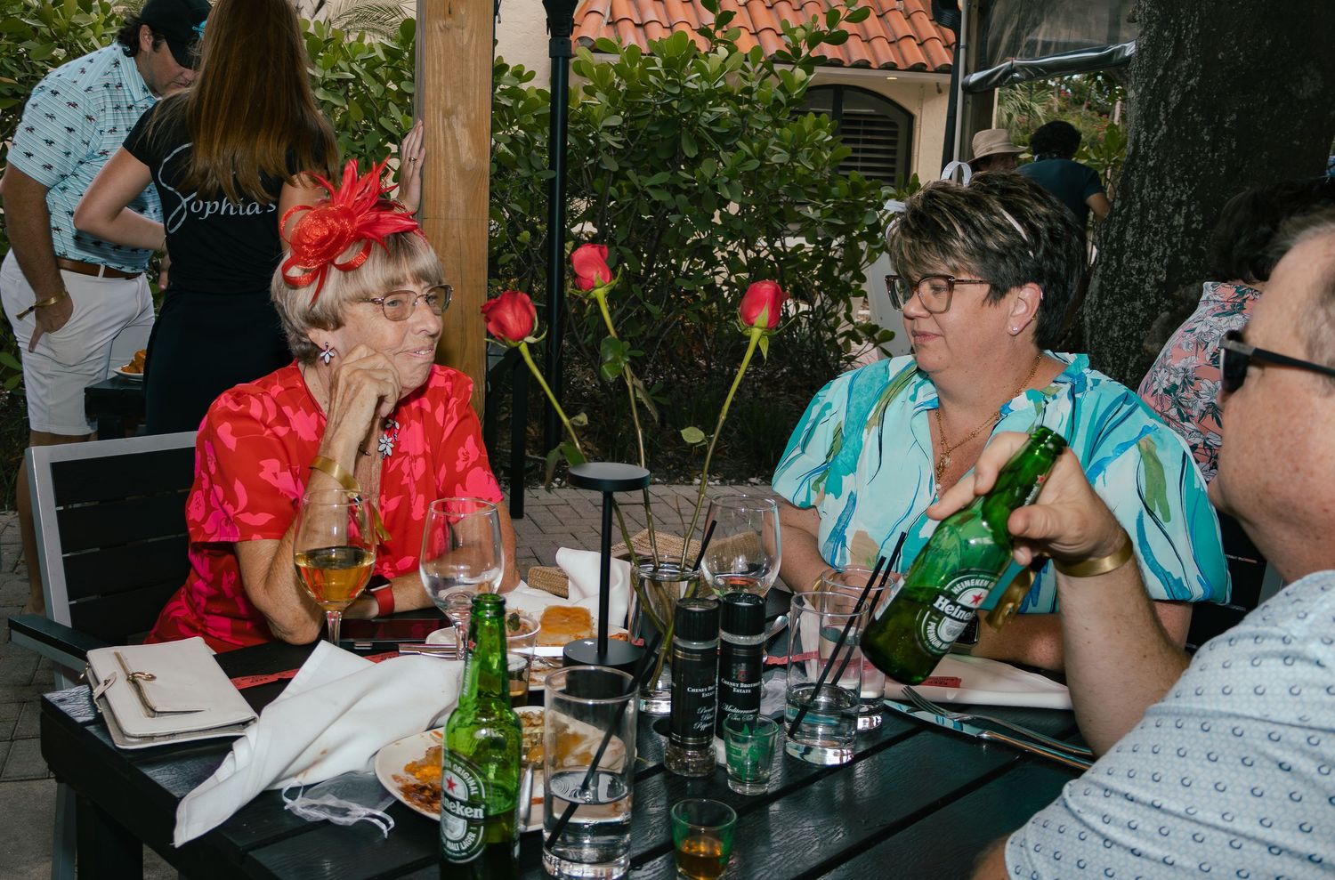Two women in colorful outfits enjoying drinks at an outdoor patio table with Heineken bottles.