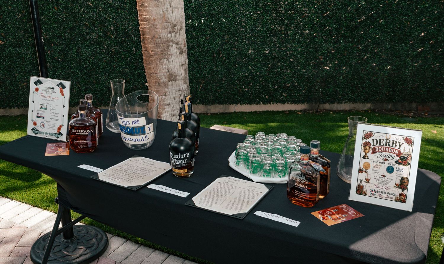 Kentucky Derby themed outdoor bar table with whiskey bottles, water, and signage on black tablecloth.