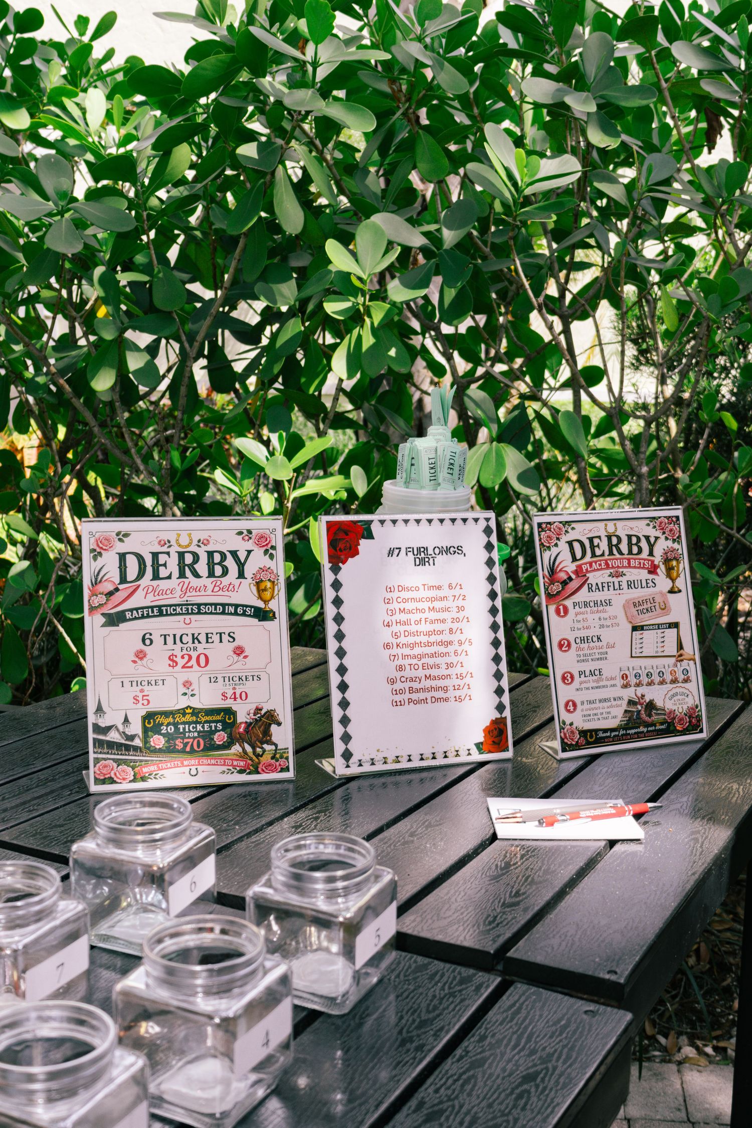 Derby raffle ticket signs displayed on a black wooden table with empty glass jars outdoors.