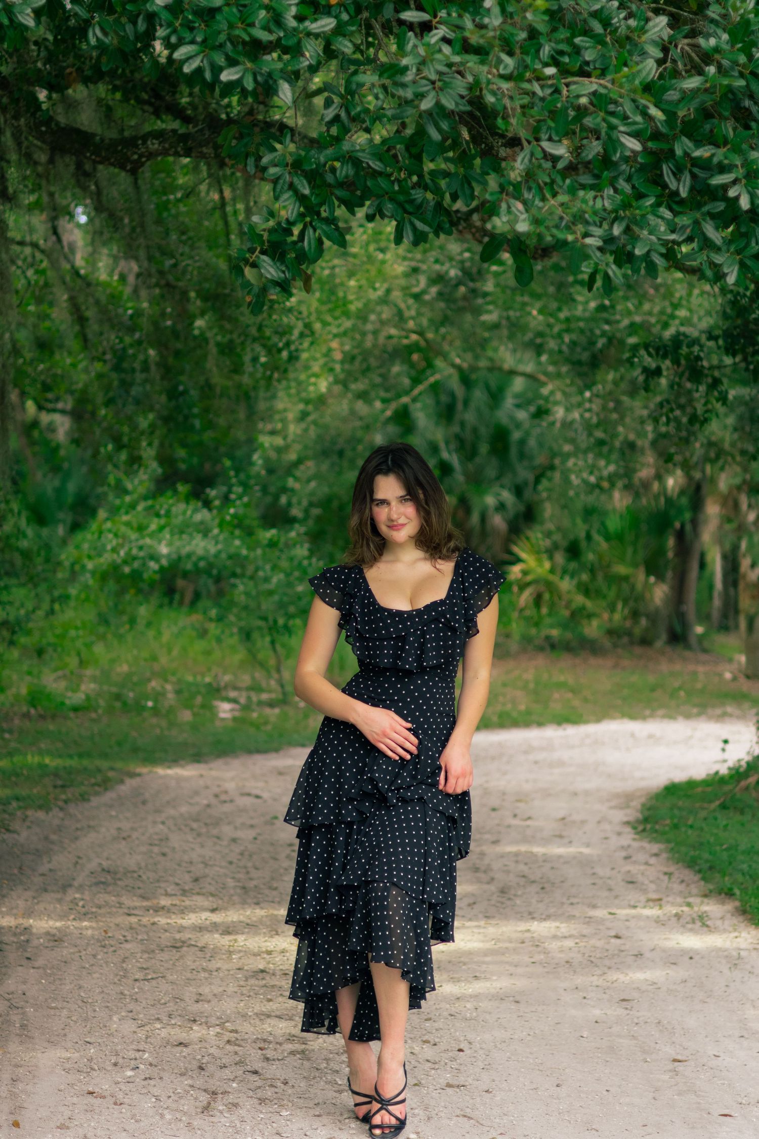 Young woman in black polka dot ruffled dress walking down a tree-lined dirt path outdoors.