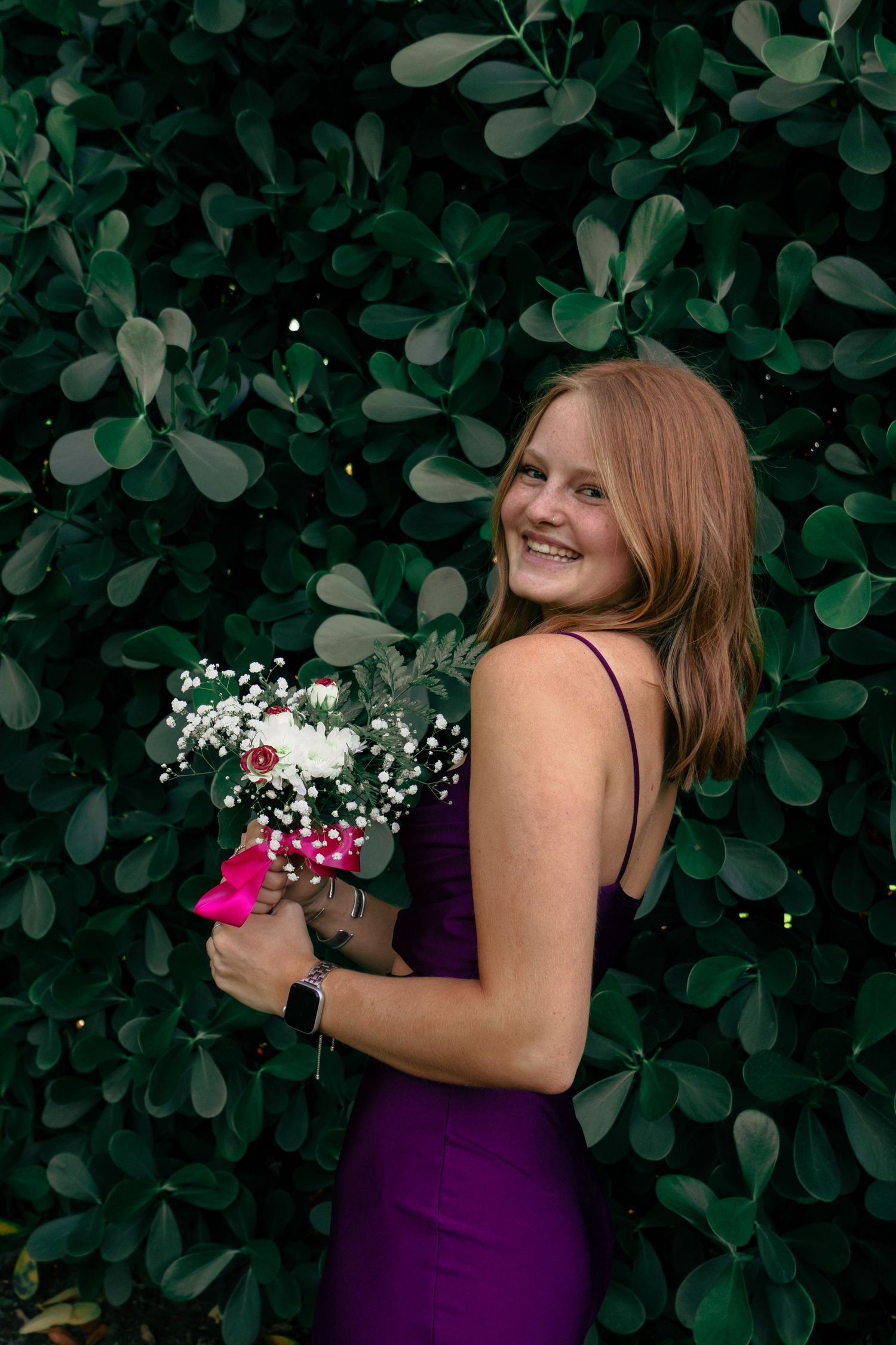 Smiling redhead in a purple spaghetti strap prom dress holding a colorful floral bouquet.