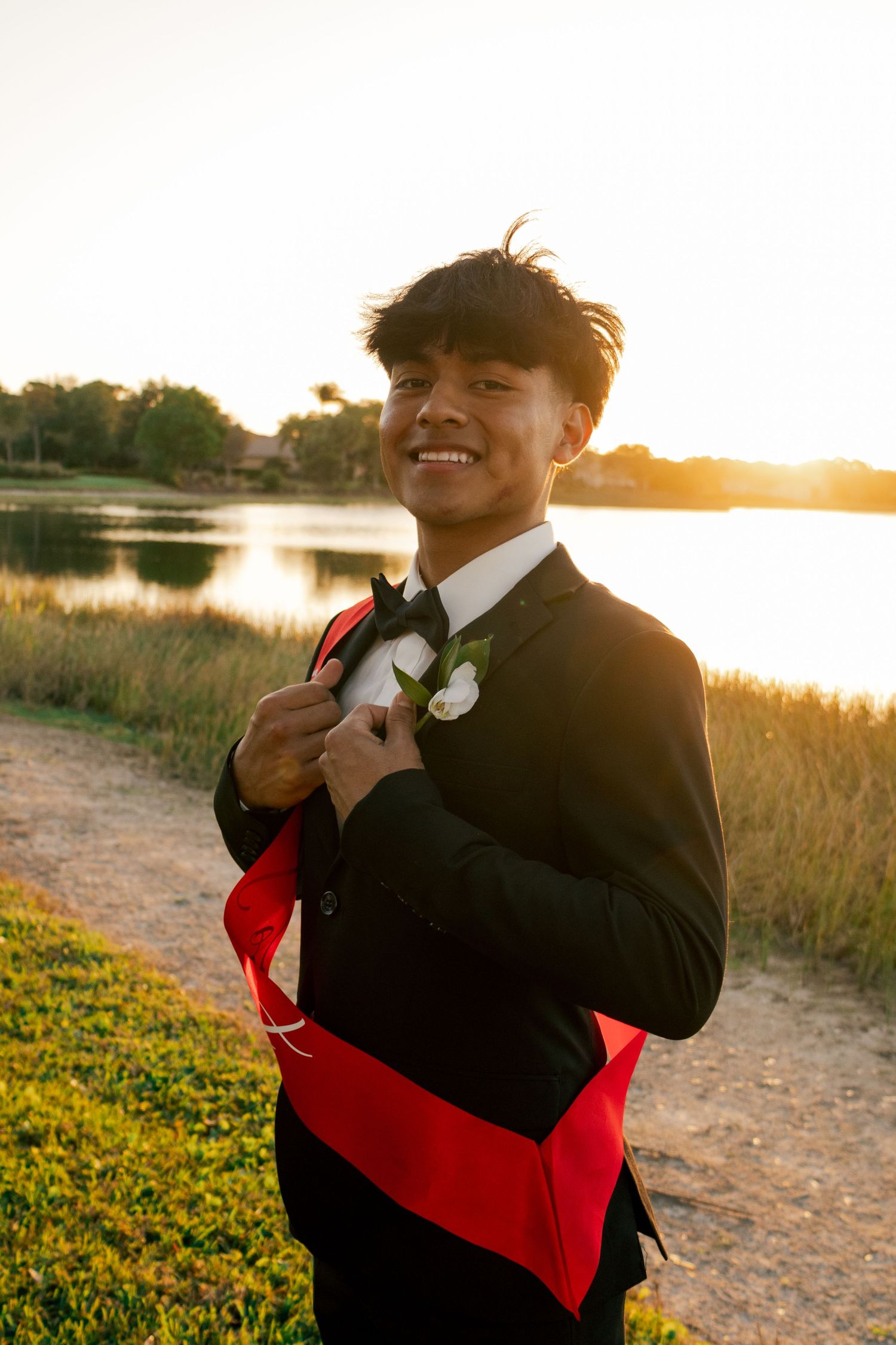 Smiling young man in black tuxedo with red sash and boutonniere at golden sunset lakeside.