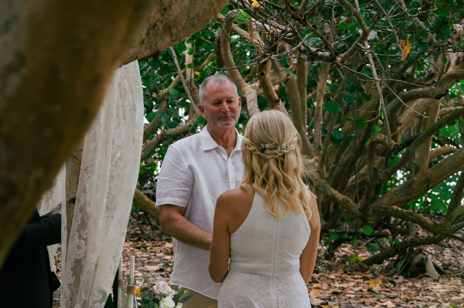 Groom smiles at bride during intimate outdoor wedding ceremony among tropical trees.