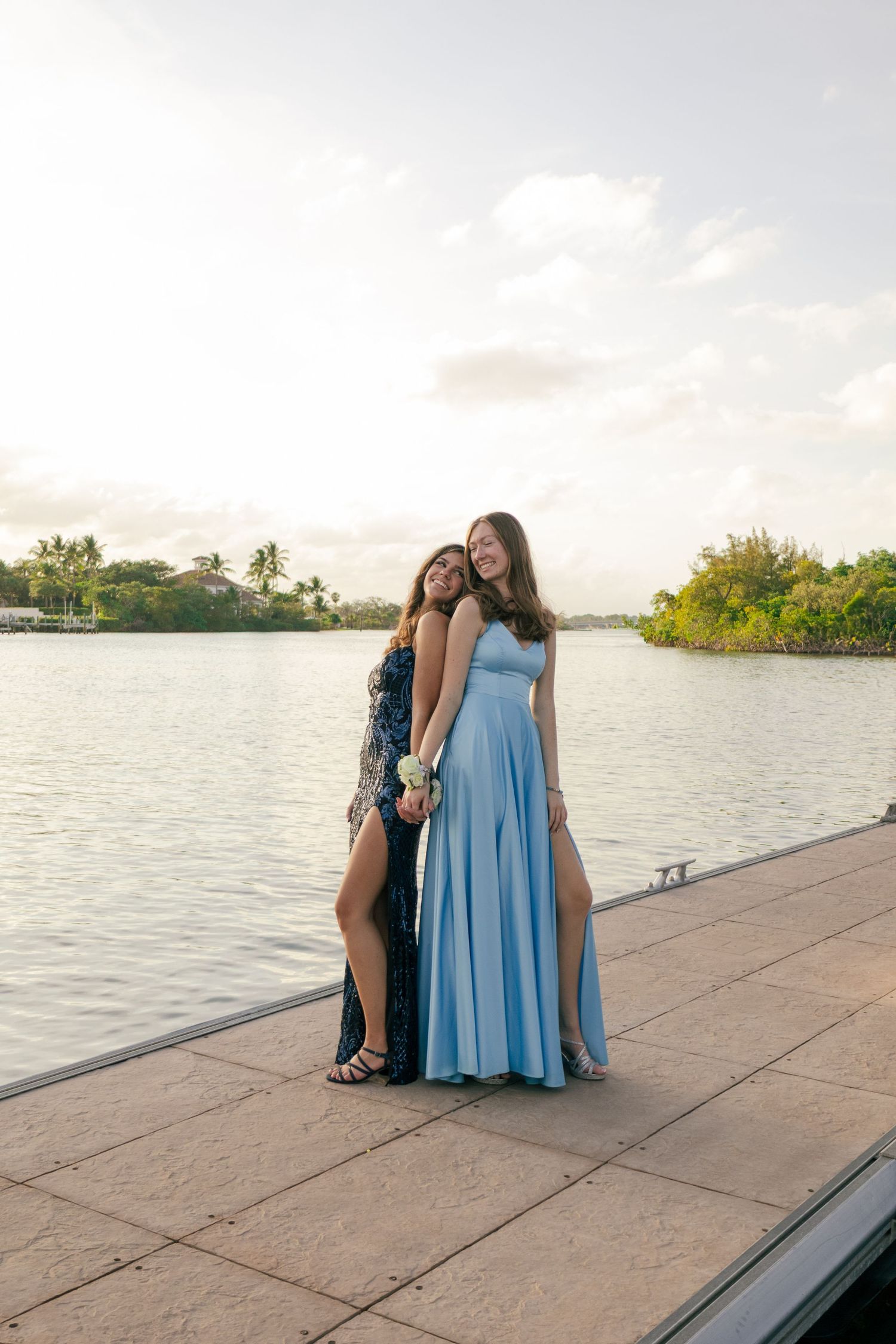 Two young women in blue formal dresses embrace while posing on a waterfront dock at dusk.