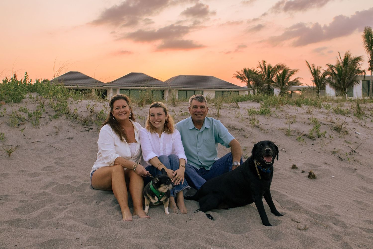 Family of three sitting on sandy beach at sunset with two dogs, tropical bungalows and palm trees in background.