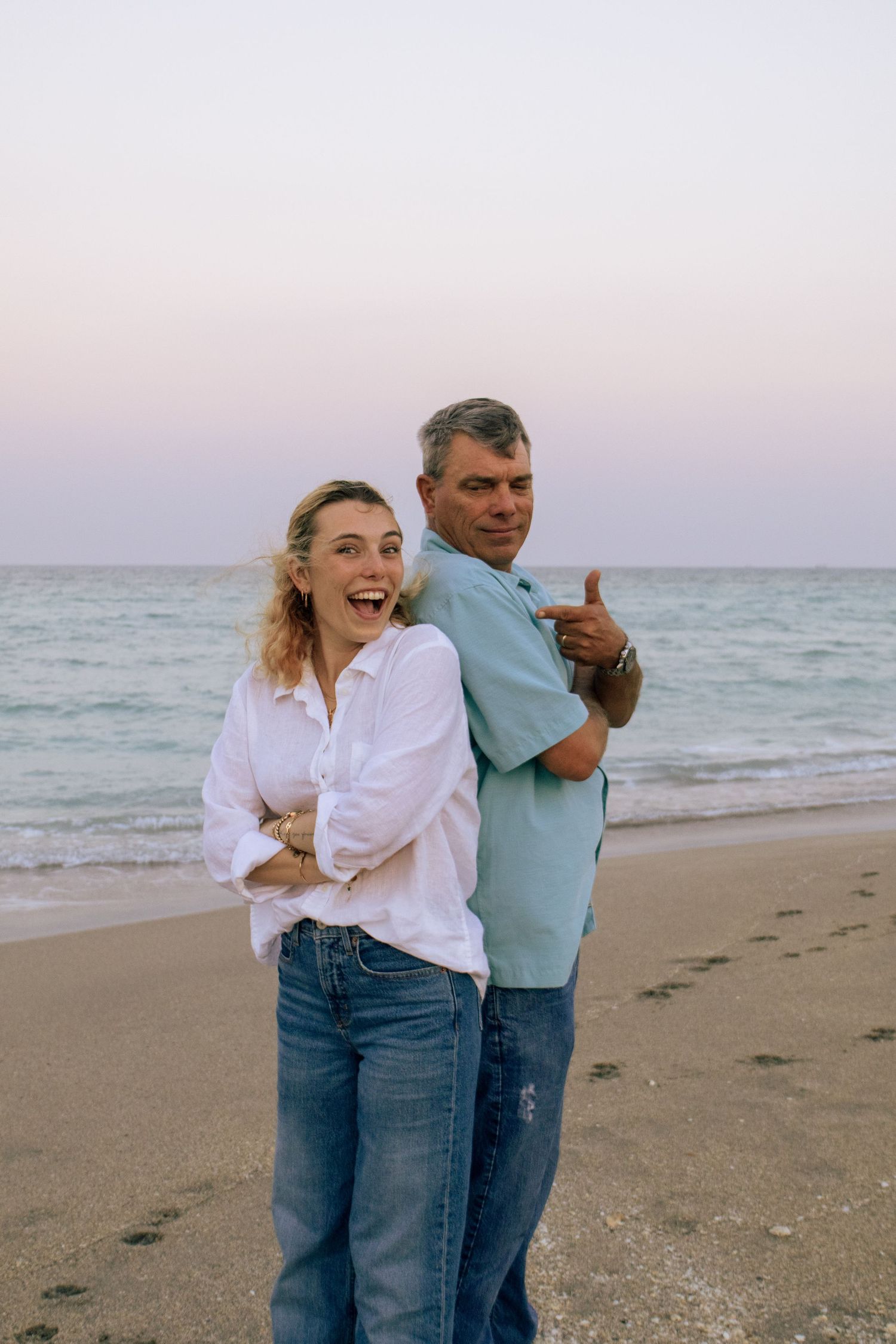 Laughing young woman and smiling man posing back-to-back on beach at twilight.