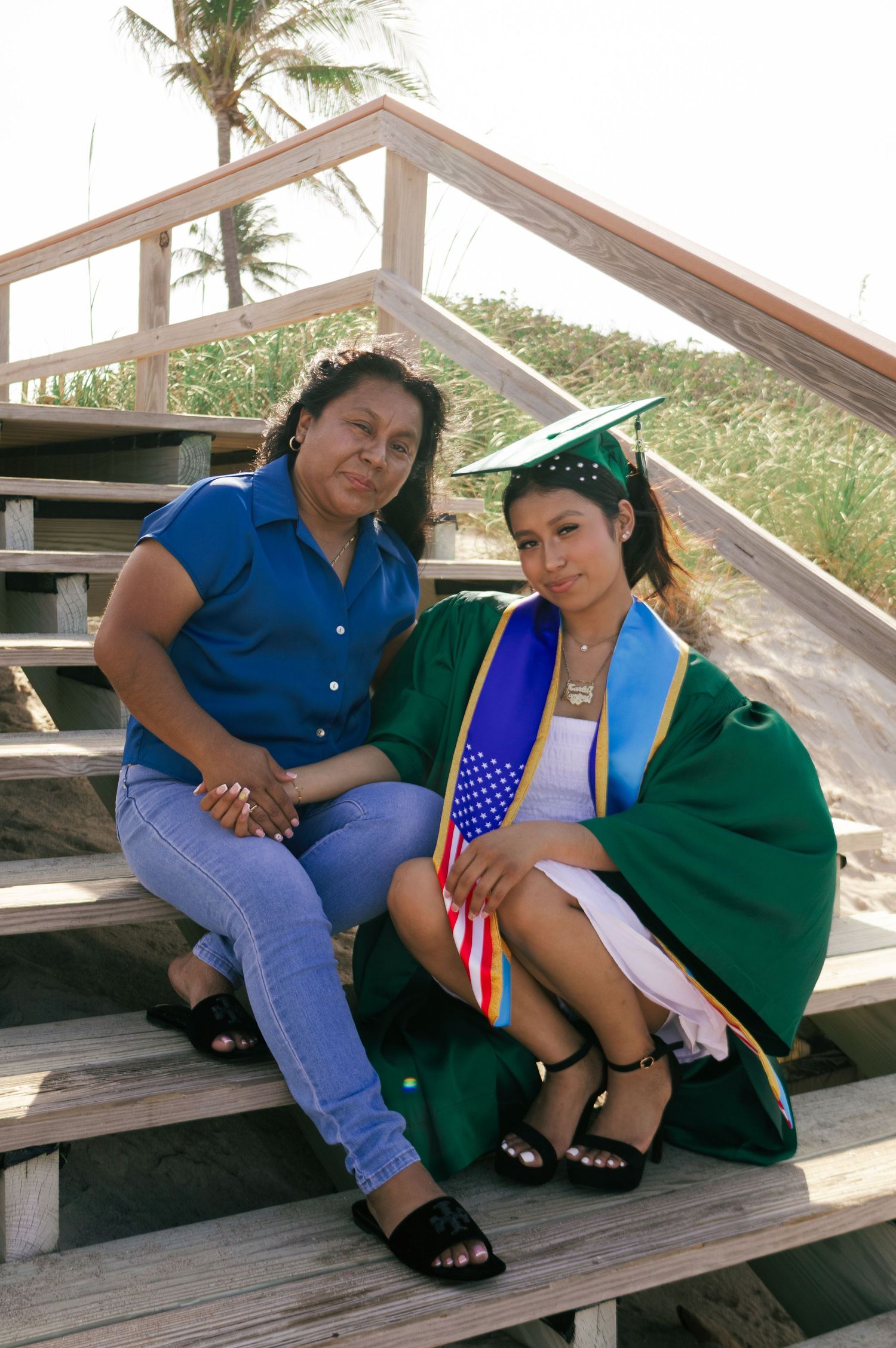 Graduate in green cap and gown sits with mother on wooden beach boardwalk stairs holding American flag.