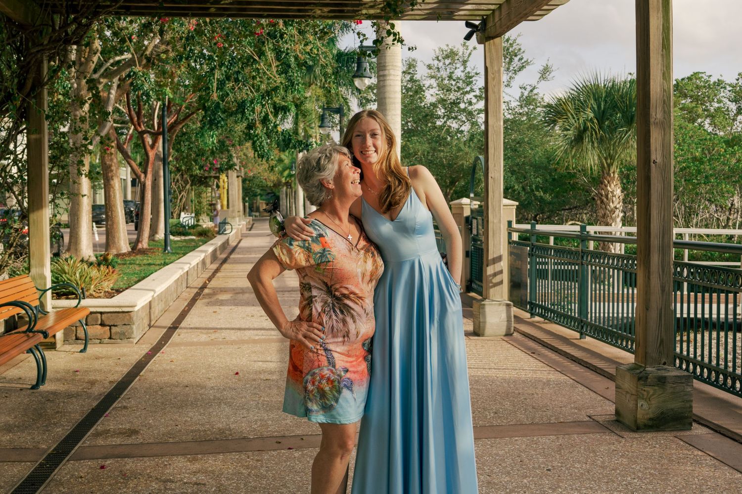 Two women in elegant dresses pose under a pergola in a lush outdoor park setting.
