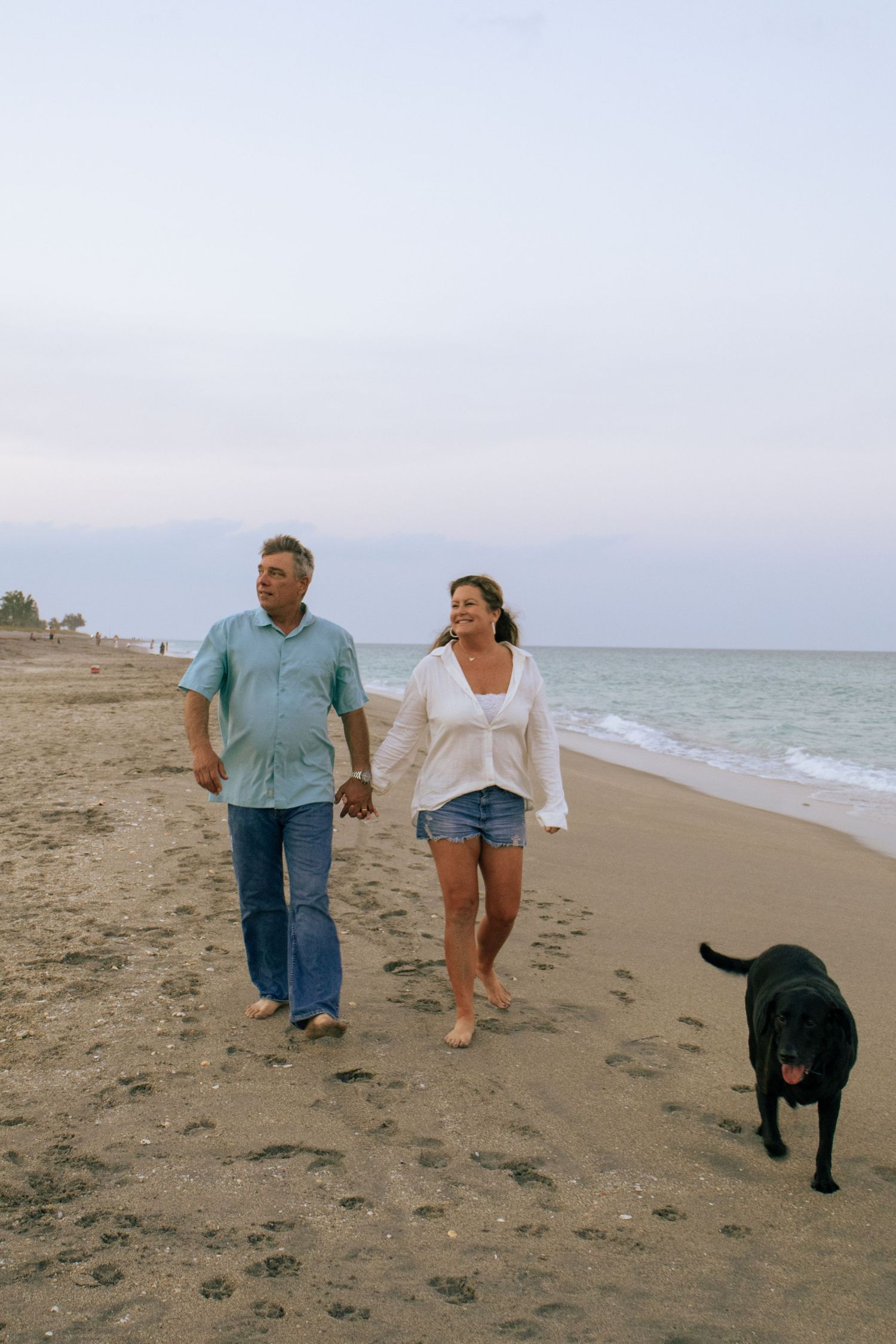 Couple holding hands walking barefoot on sandy beach with black Labrador dog at dusk.