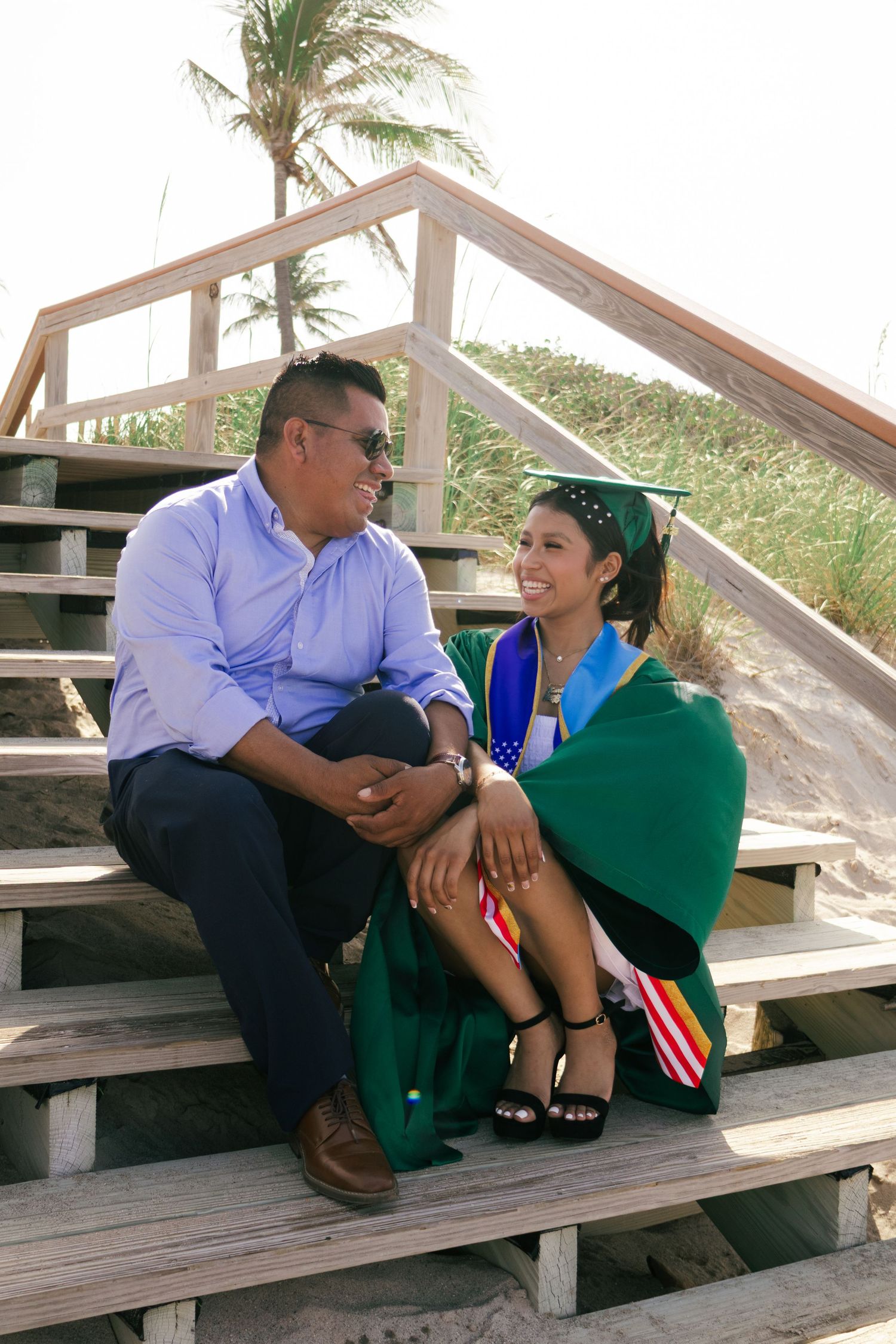 Graduate in green cap and gown sits with father on sunny beach boardwalk stairs smiling at each other.