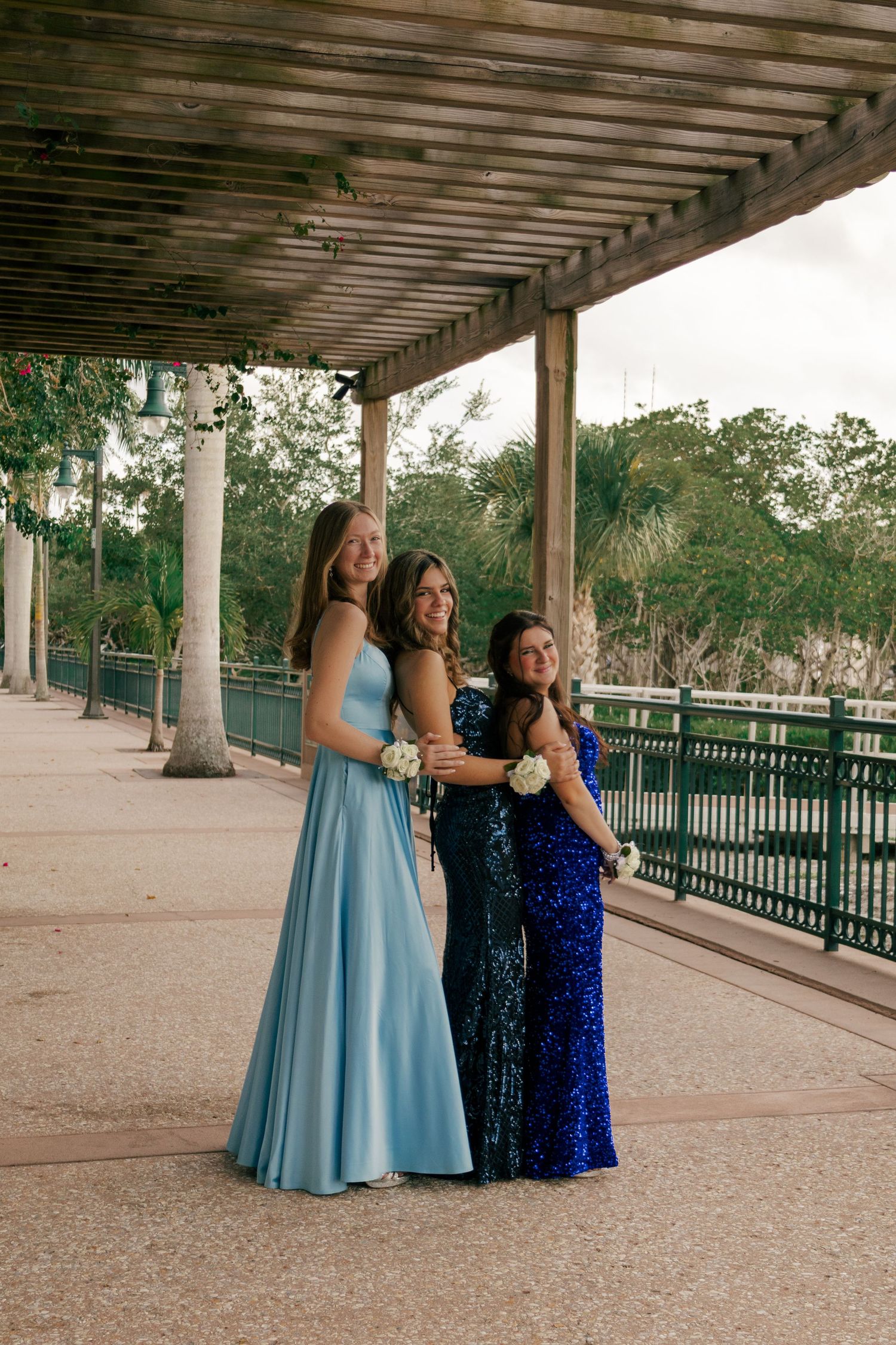 Three young women in blue formal gowns pose together under a wooden pergola near a waterway.