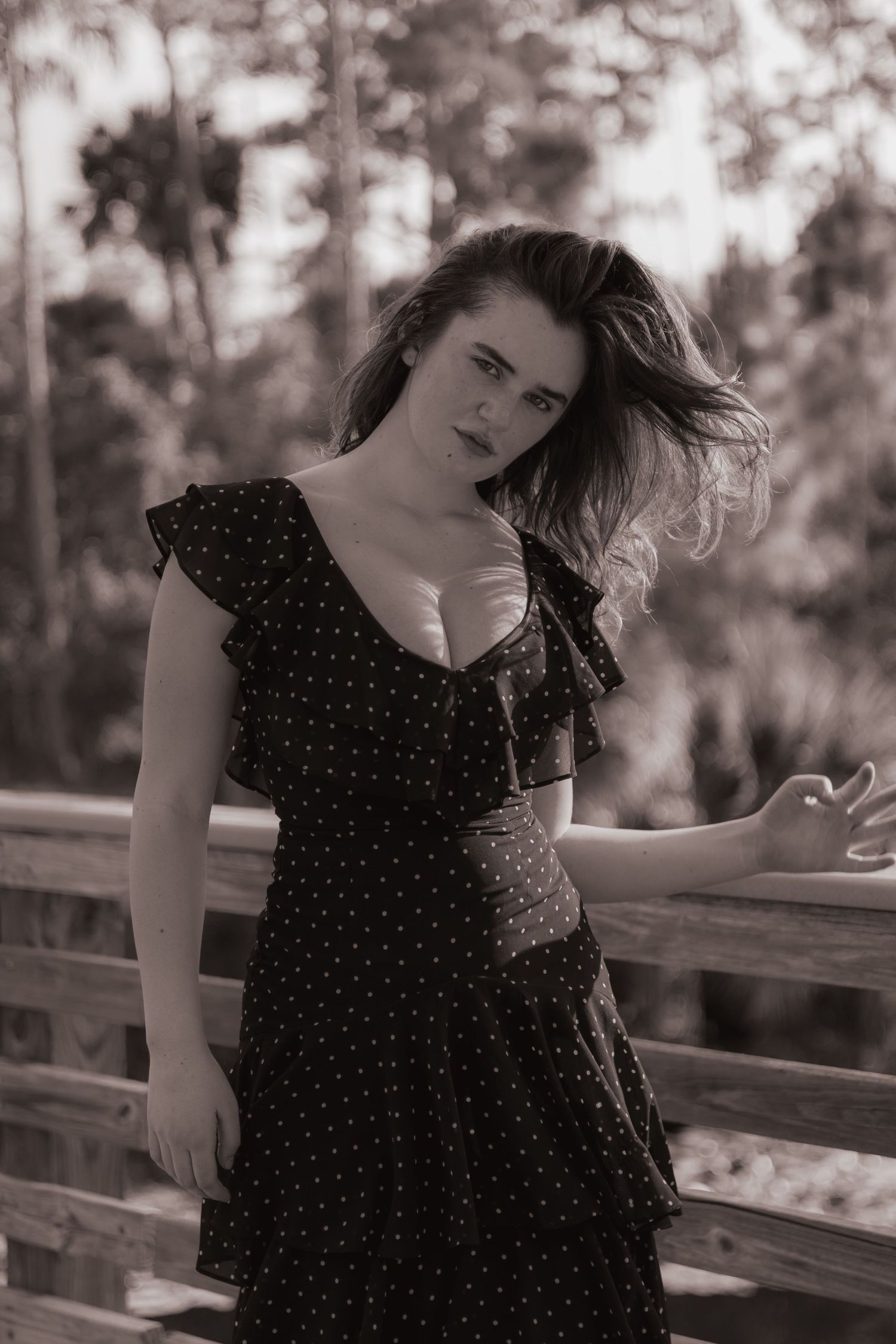 Black and white portrait of woman in polka dot dress posing on wooden bridge with windswept hair.