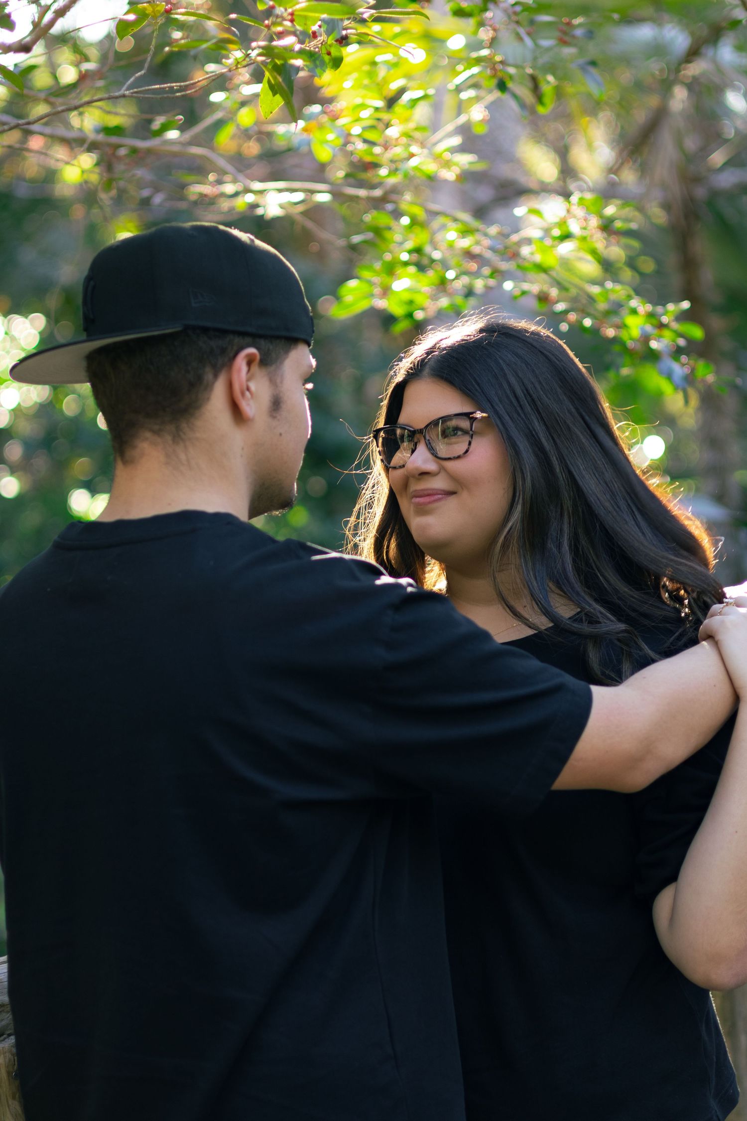 Young couple in black outfits facing each other and embracing near wooden fence with sunlit green trees.