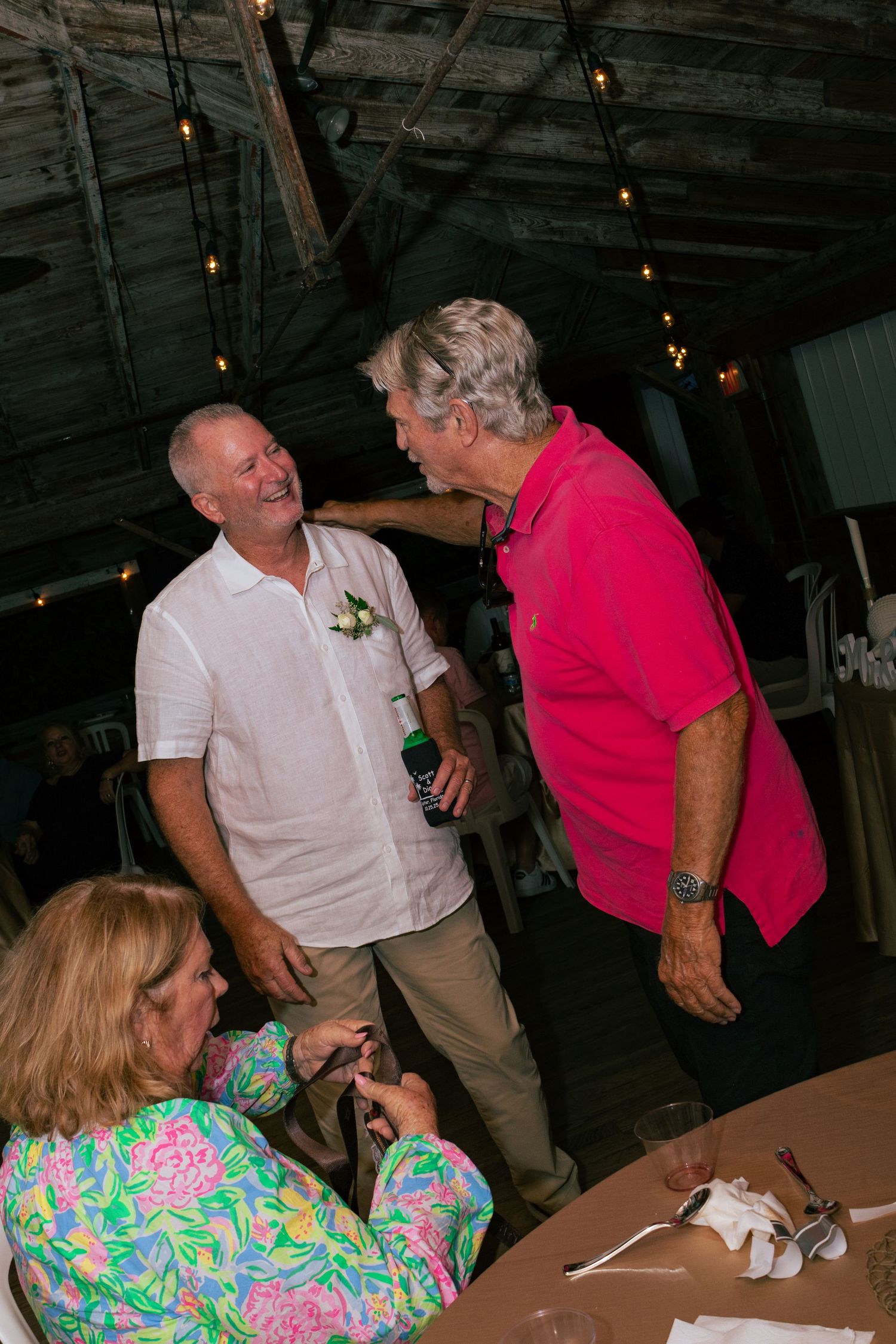 Groom in white shirt laughs with guest in pink polo at wedding reception.