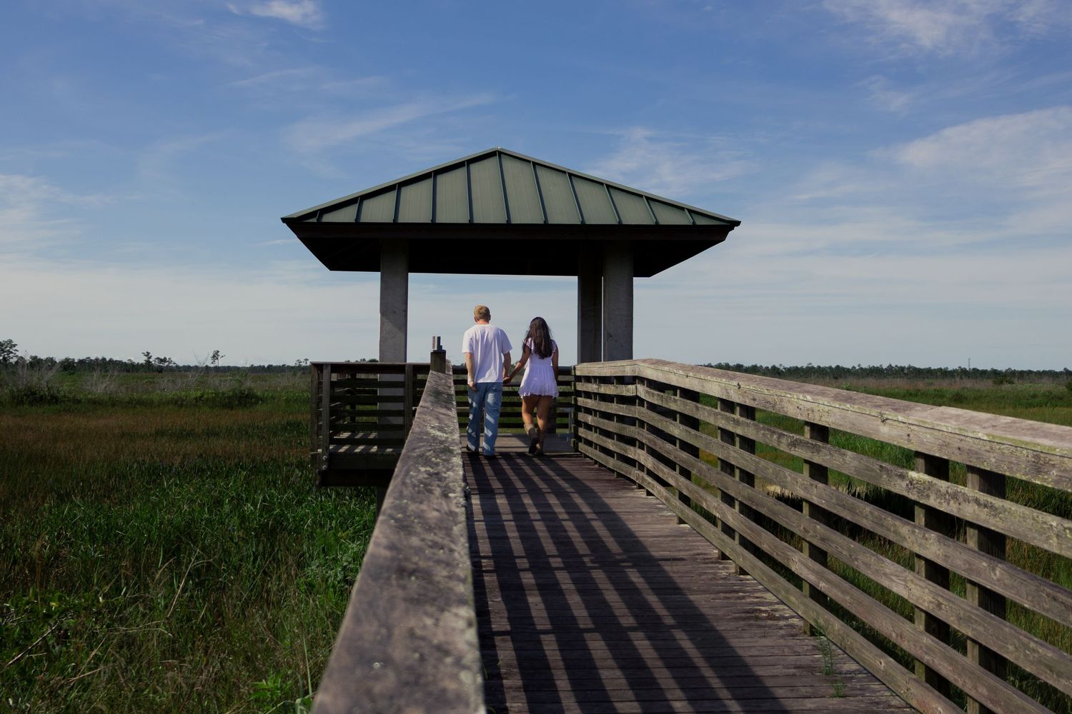 Couple walking hand in hand toward a covered gazebo on a wooden boardwalk over marshlands.