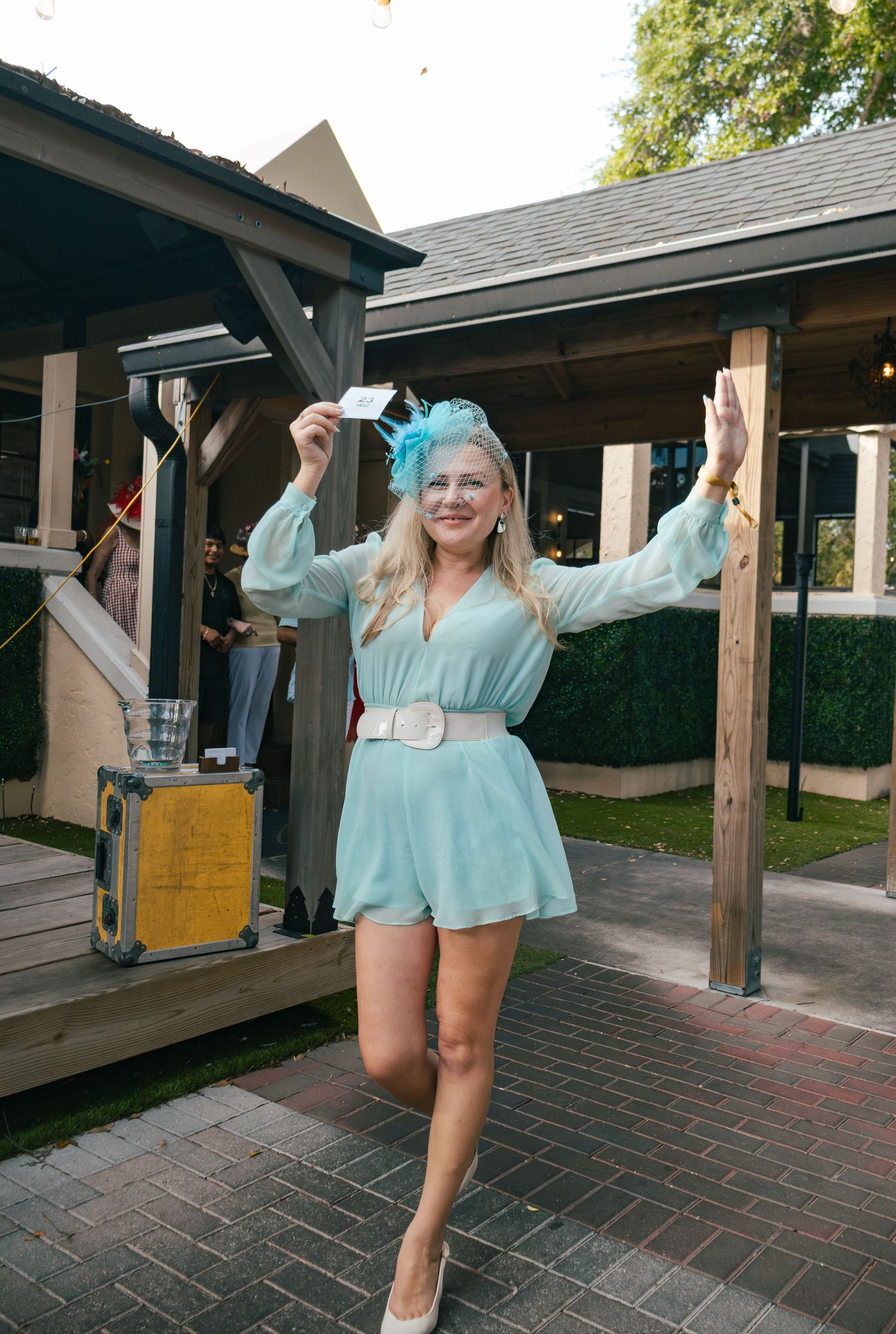Blonde woman in mint green romper with blue fascinator celebrates at Derby party.