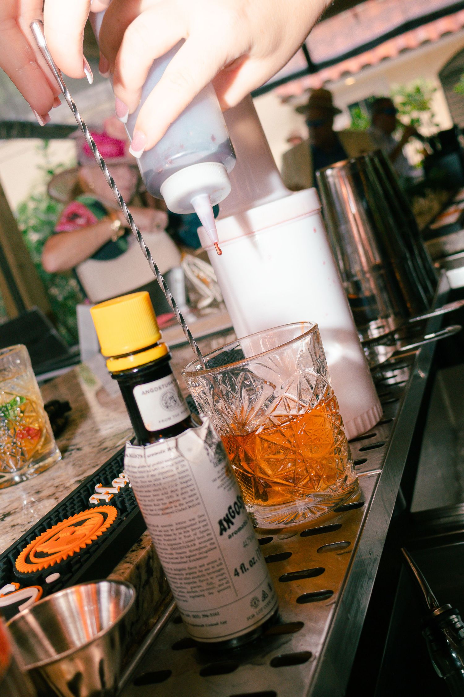Bartender pouring a cocktail into a crystal glass with Angostura bitters and bar tools on a marble counter.