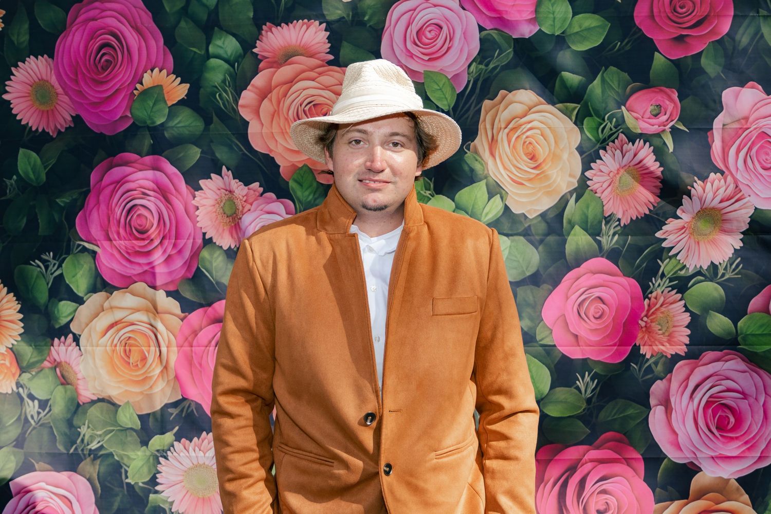 Man in an orange blazer and white hat smiling in front of a colorful floral rose backdrop.