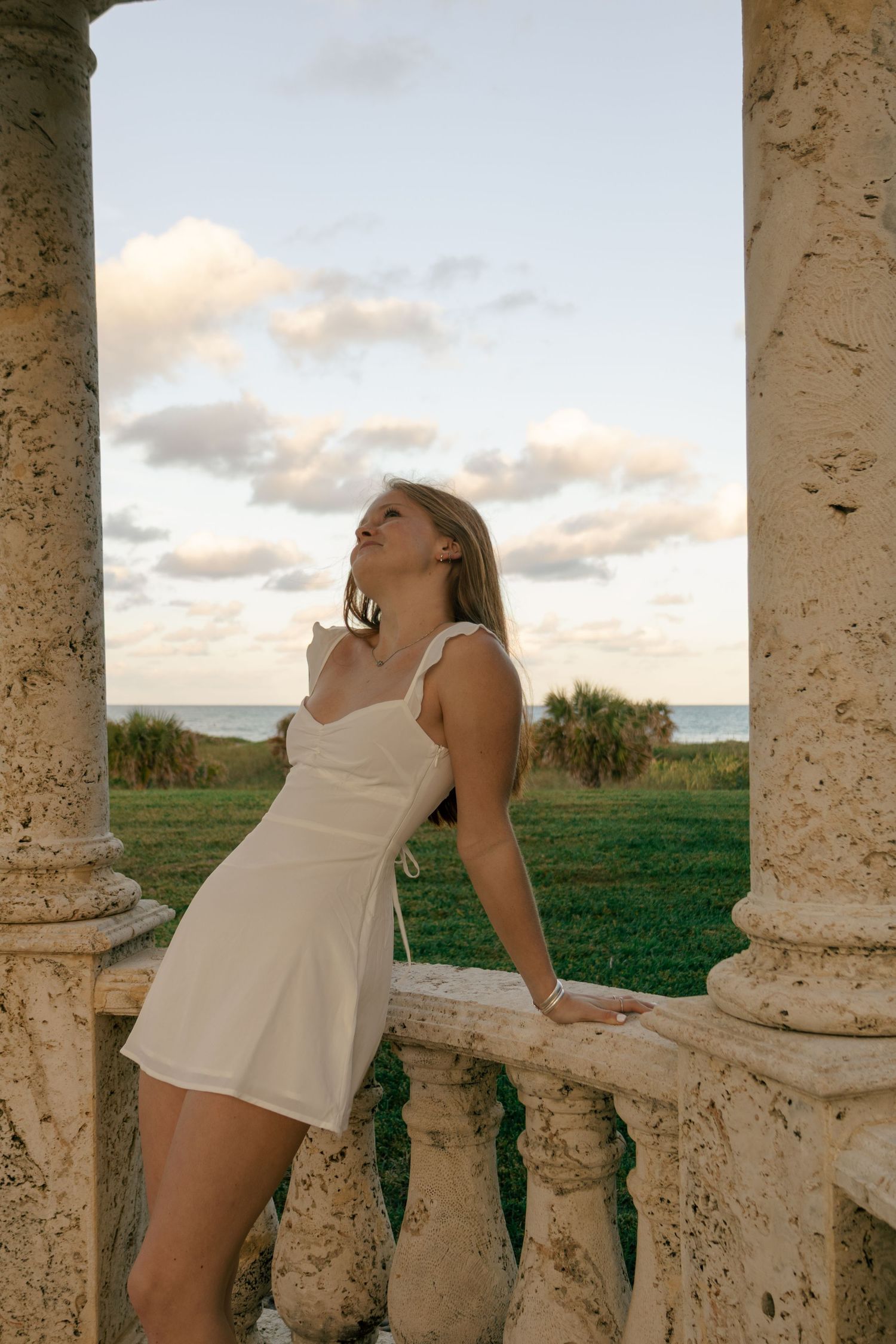 Young woman in white dress posing between stone columns at sunset near the ocean.