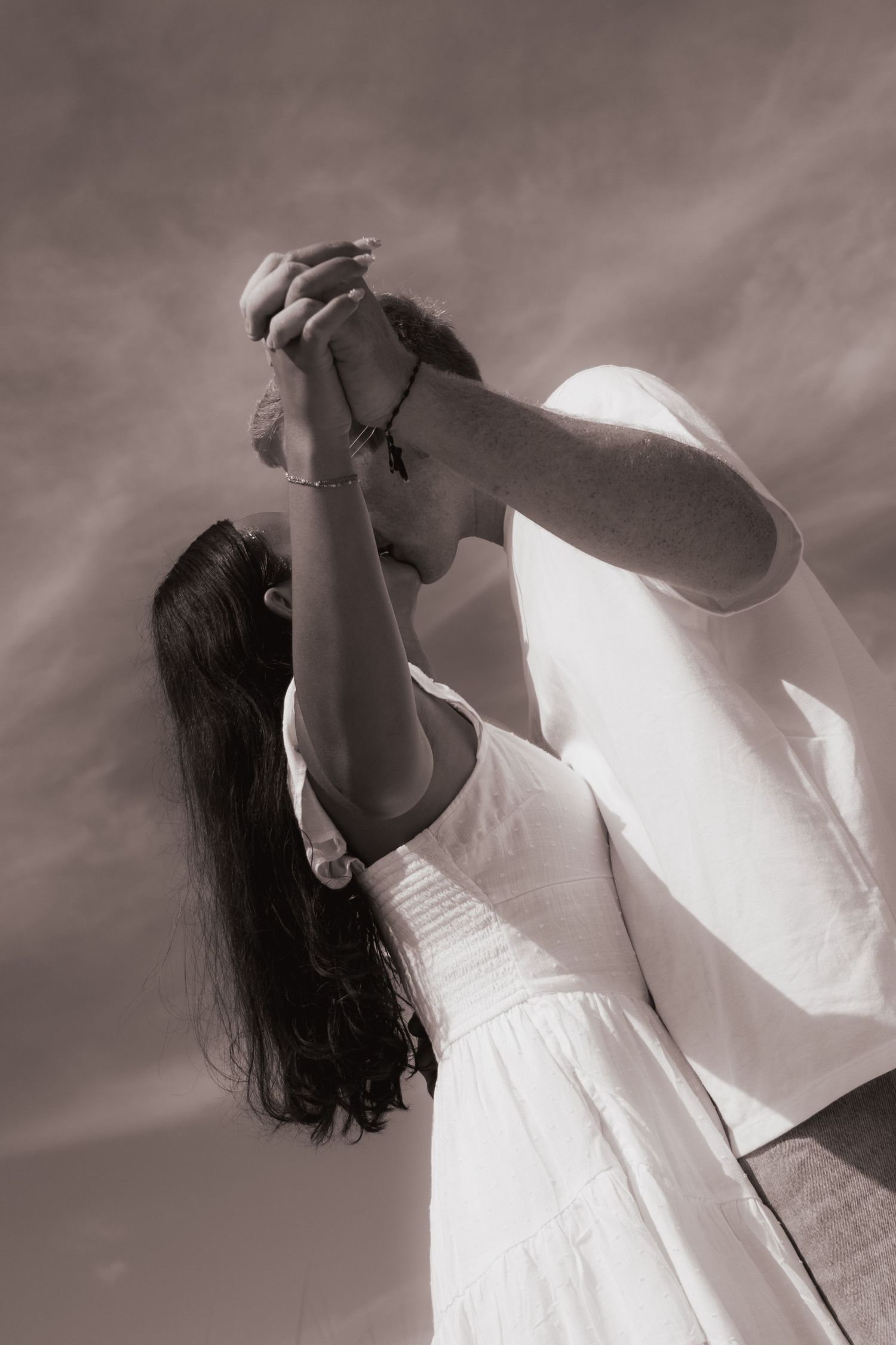 Black and white romantic photo of a couple kissing with raised hands against a cloudy sky.