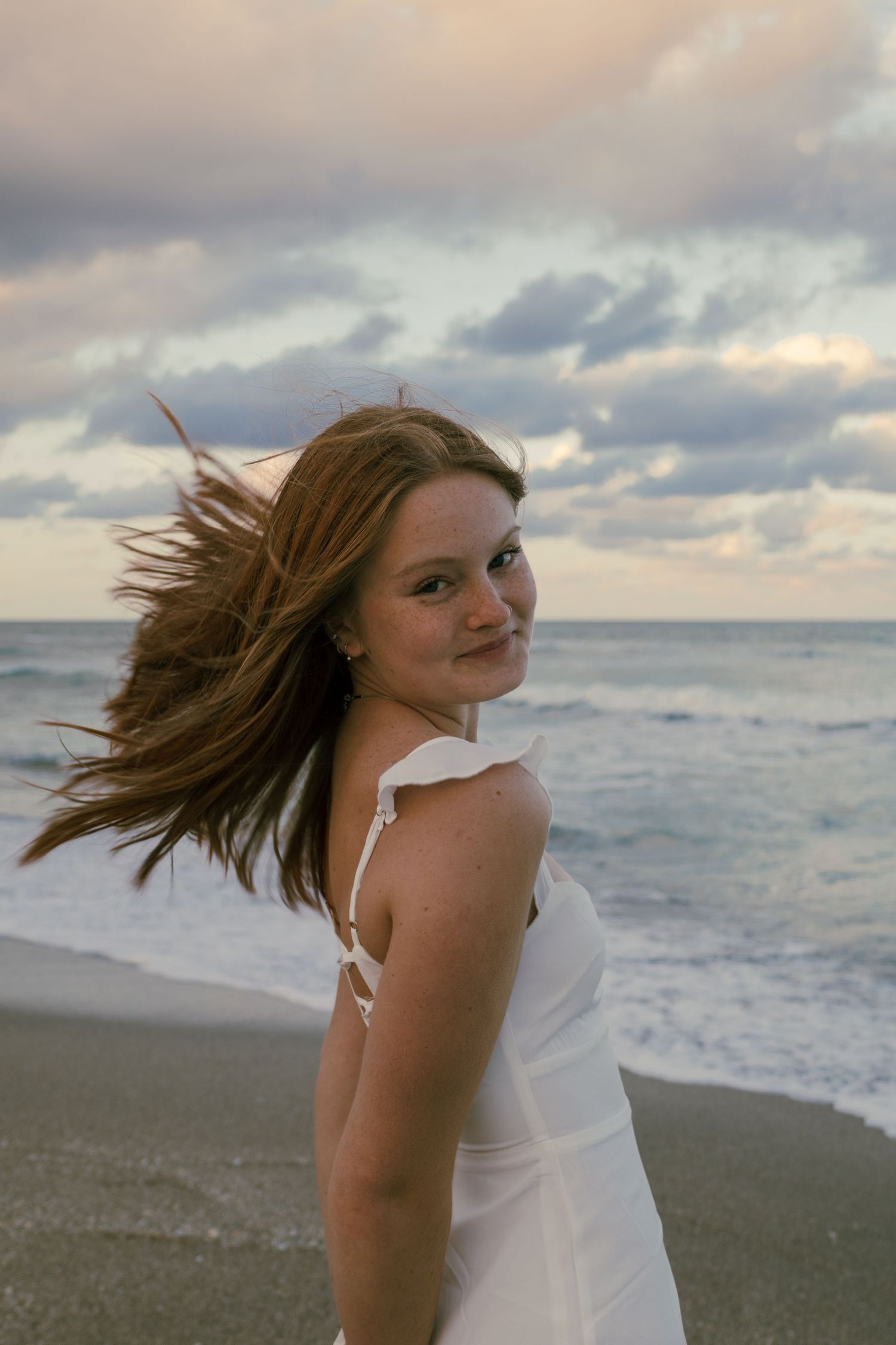 Young woman in white dress laughing with hair blowing in wind on beach at sunset with ocean waves.