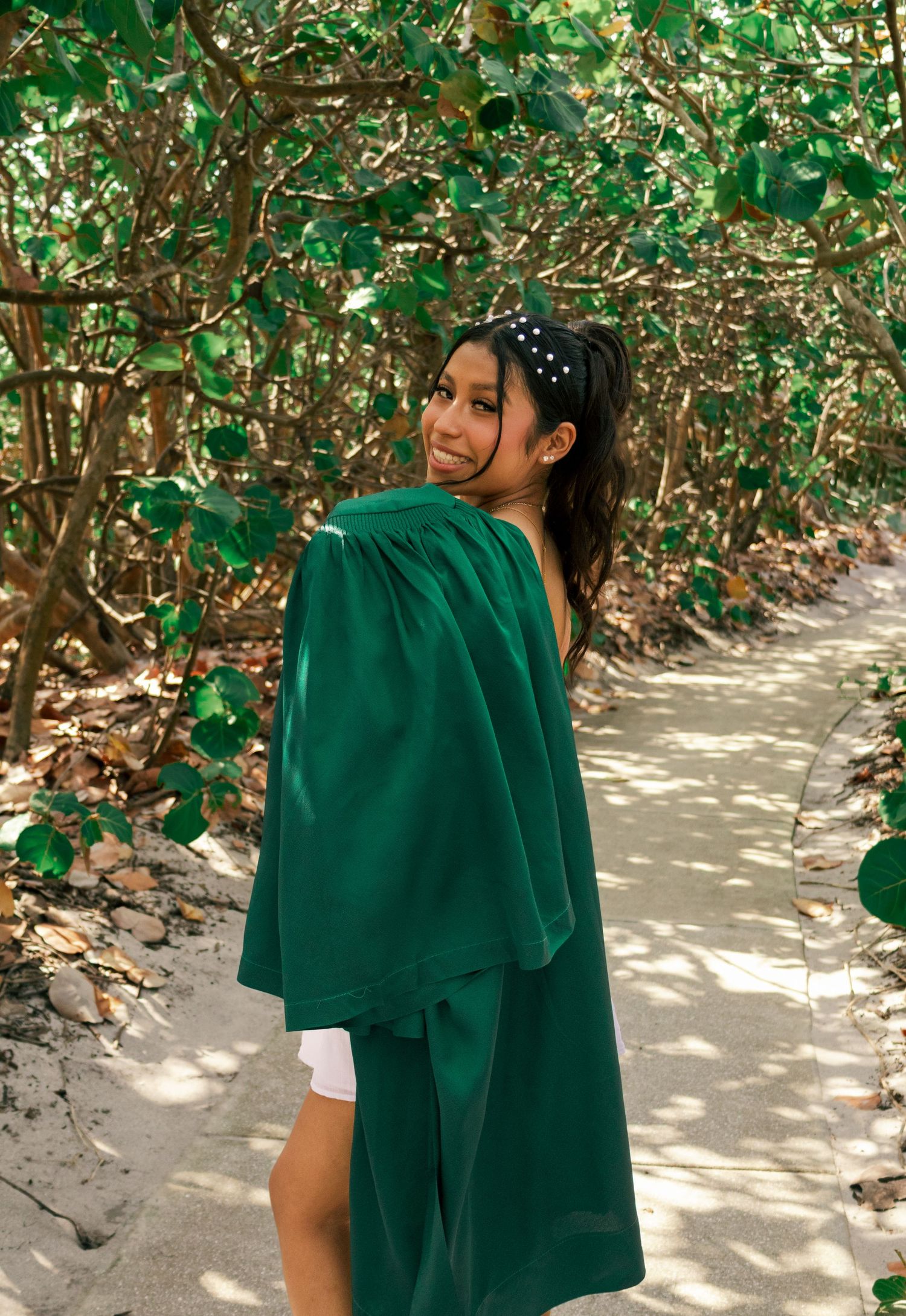 Graduate in green commencement gown walking sandy path through tropical trees, glancing back over shoulder.