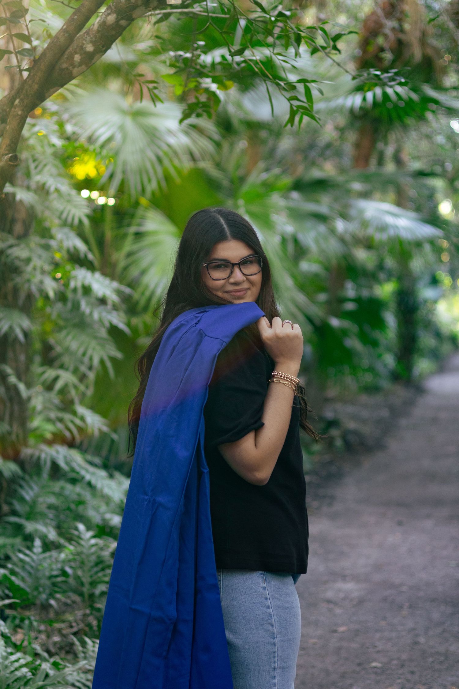 Graduate glancing back while holding blue graduation gown on shoulder in lush tropical setting.