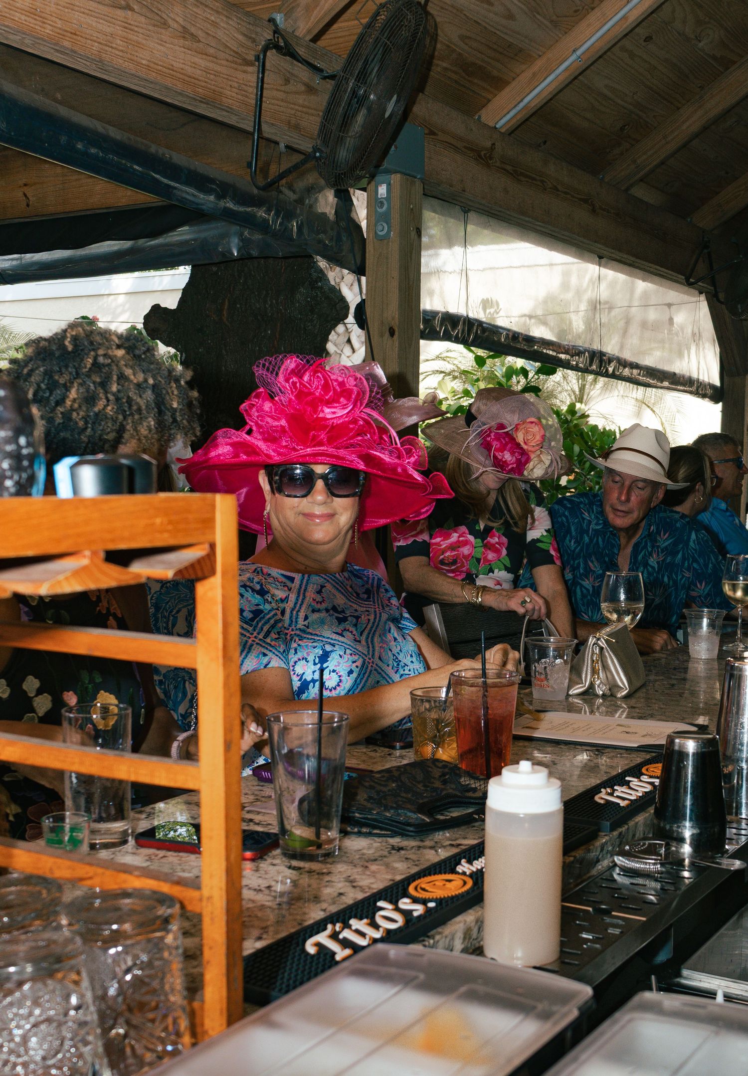 Woman in vibrant pink floral hat and sunglasses sits at a rustic outdoor bar during a festive event.