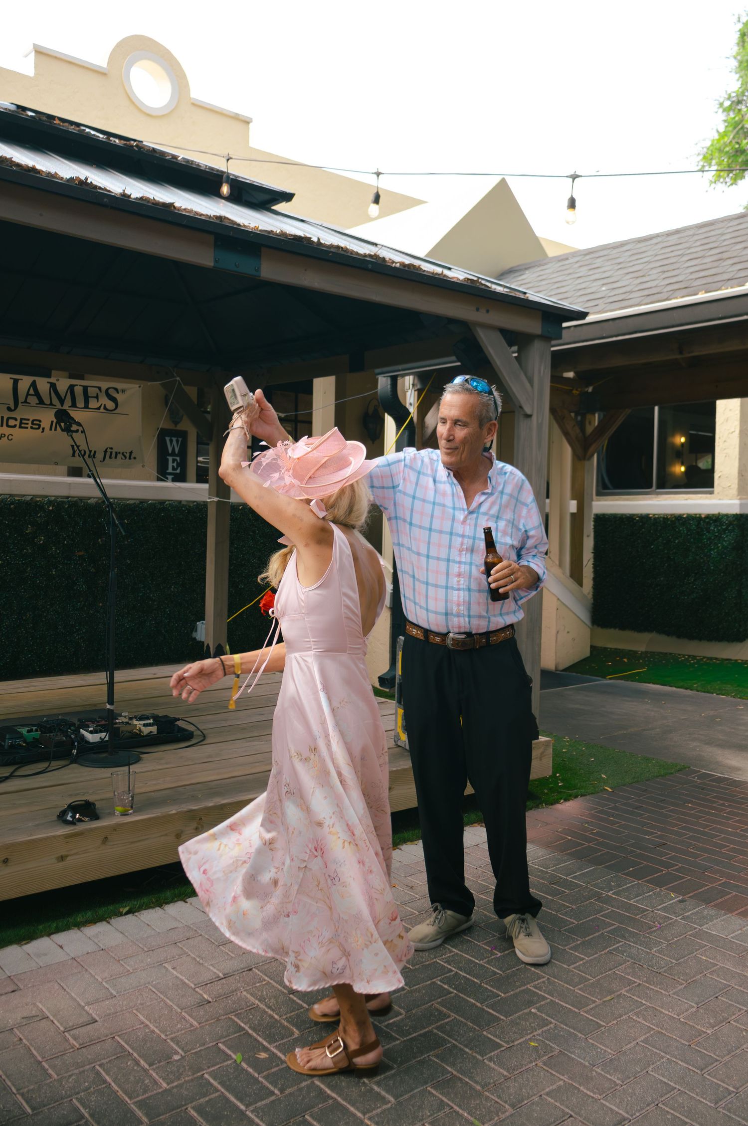 Couple at outdoor party, woman in pink dress twirling while man holds beer bottle nearby.