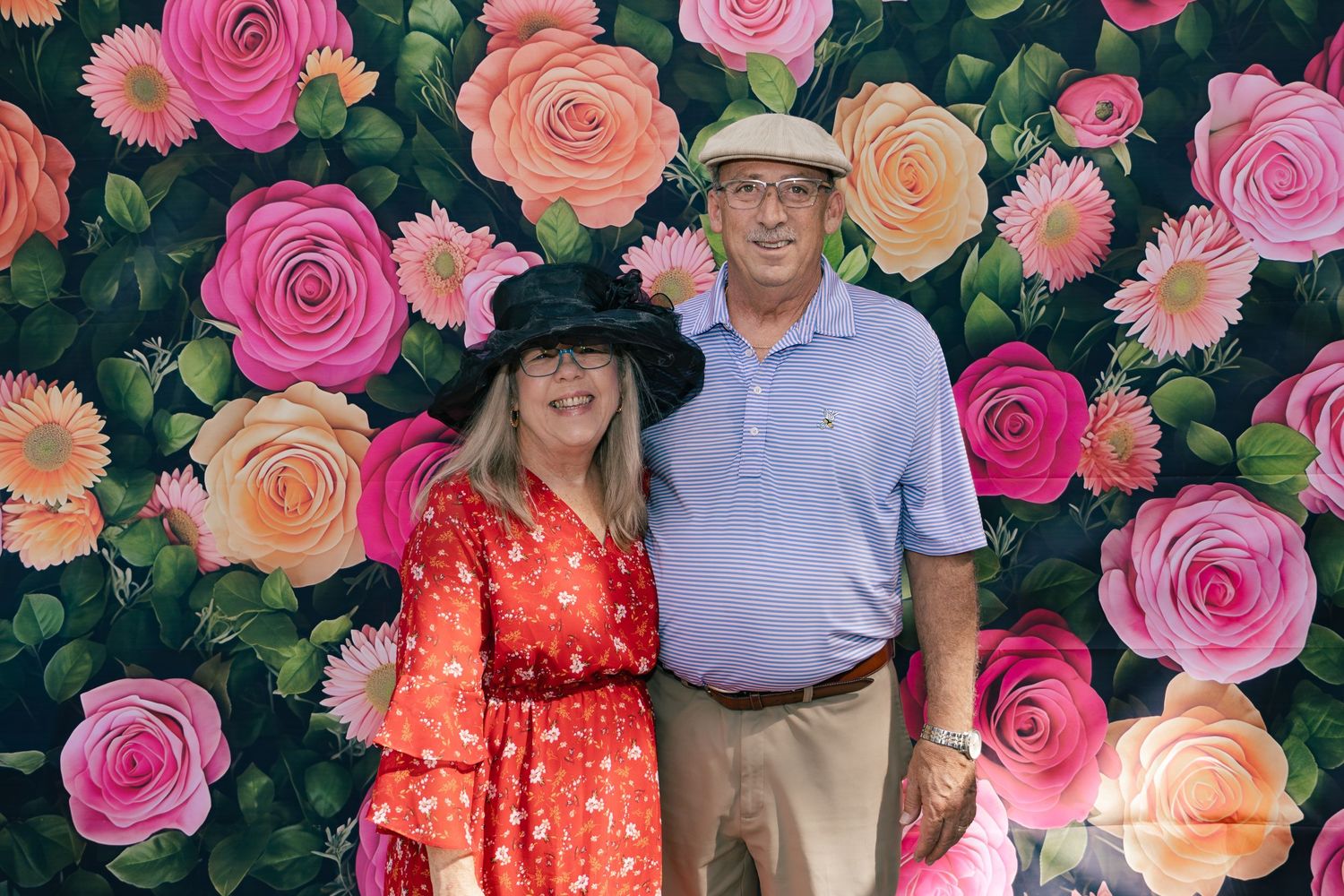 Couple wearing hats smiling together in front of a bright colorful floral backdrop with pink roses.