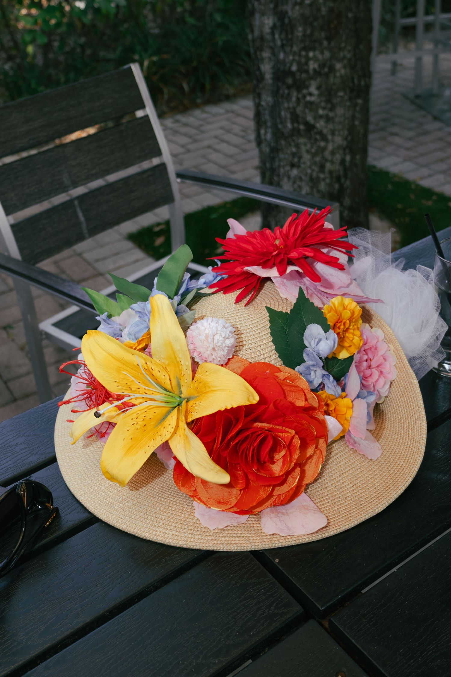 Straw hat decorated with colorful artificial flowers including yellow lily and red rose on outdoor table.