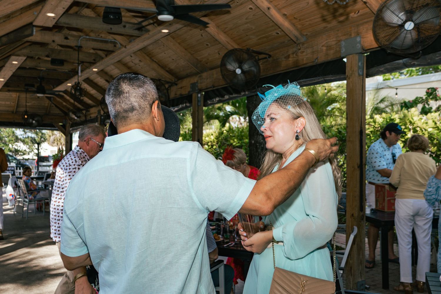Guests in Kentucky Derby attire interact under covered outdoor venue with ceiling fans.