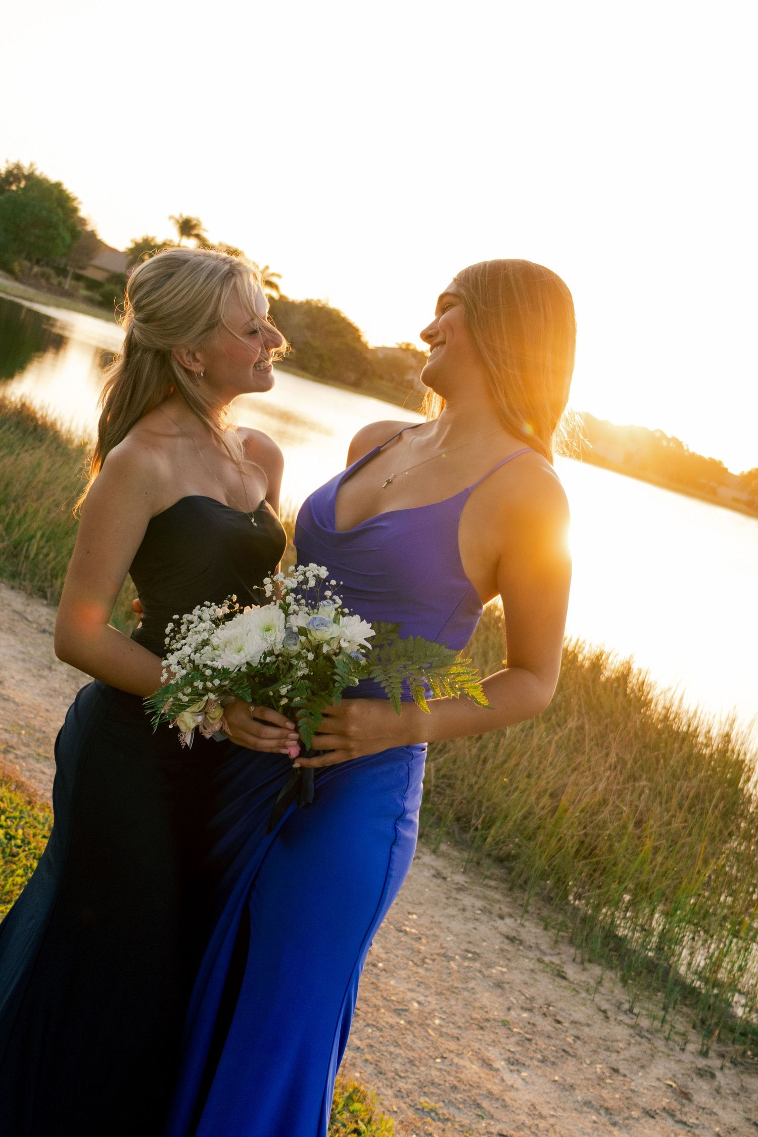 Two women in black and blue formal gowns share a joyful moment at golden sunset lakeside.