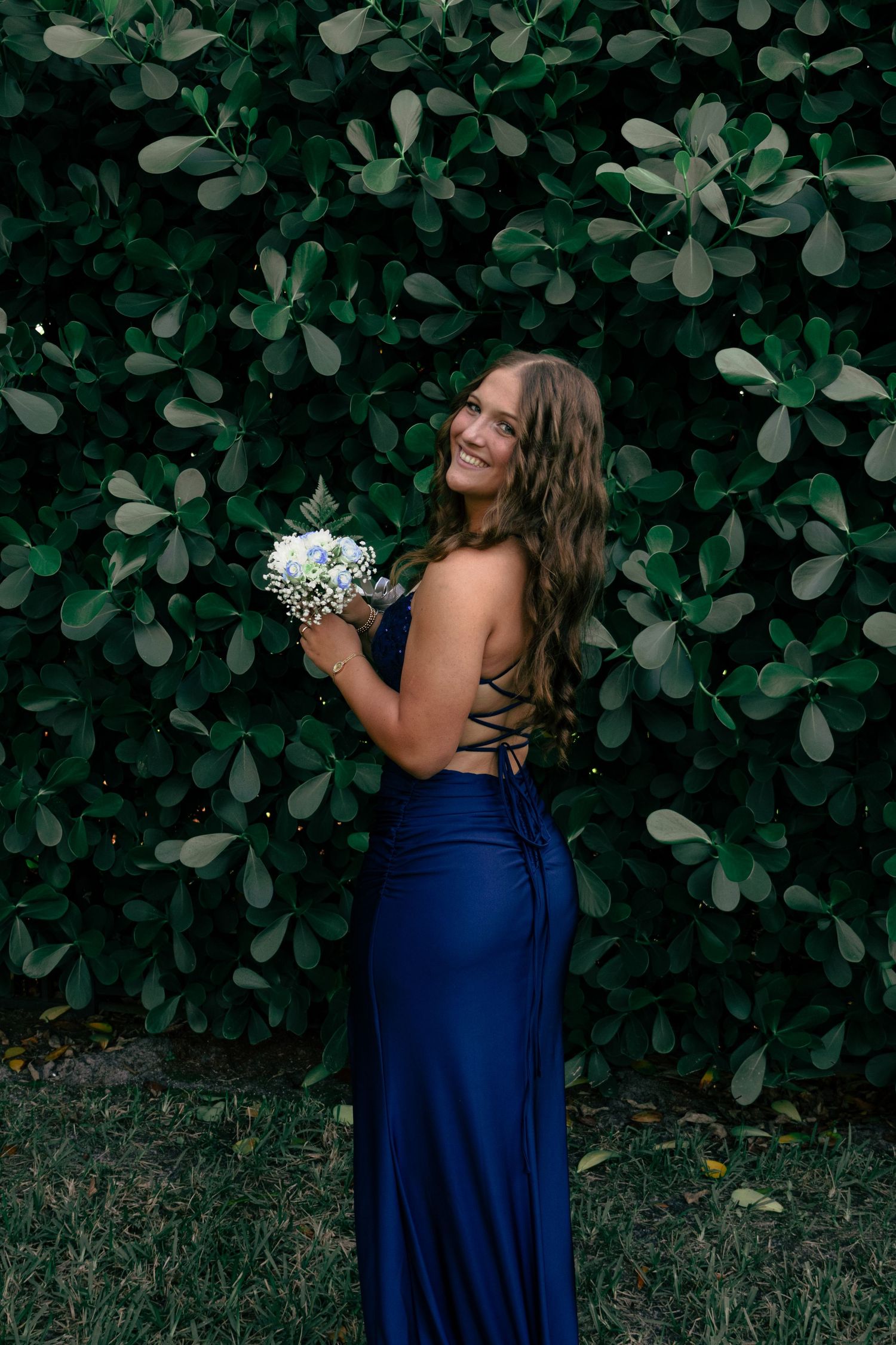 Smiling brunette in a navy backless prom gown holding a white bouquet by a green hedge.