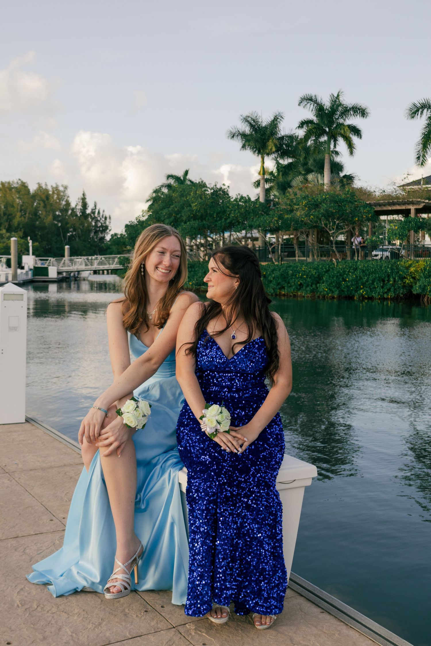 Two teenage girls in blue prom dresses sit on a waterfront dock surrounded by palm trees.