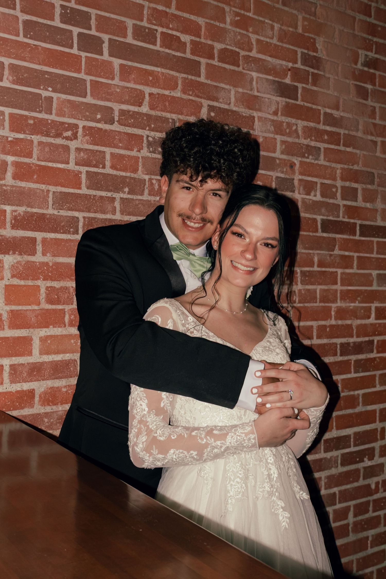 Newlywed couple embraces and smiles in front of brick wall at wedding reception.