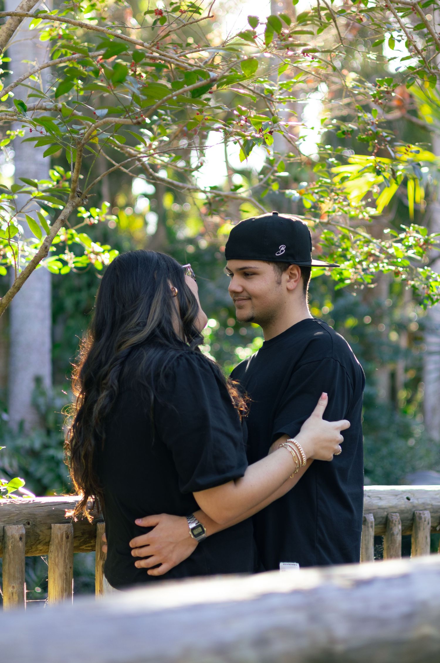 Young couple in matching black outfits embracing and smiling at each other on a wooden bridge outdoors.