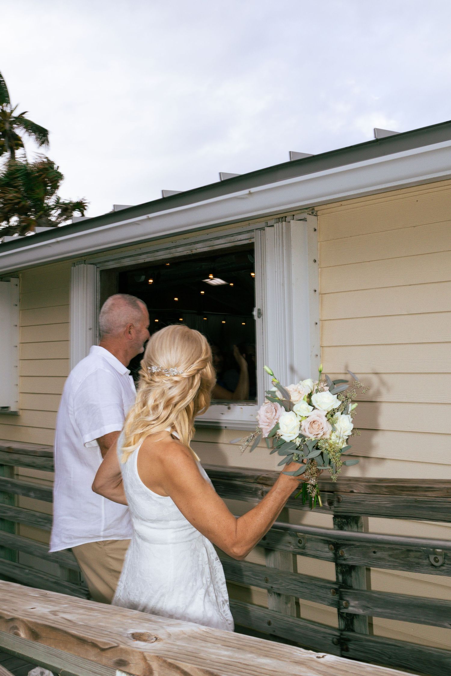 Bride holding bouquet of white roses stands with groom on wooden deck.