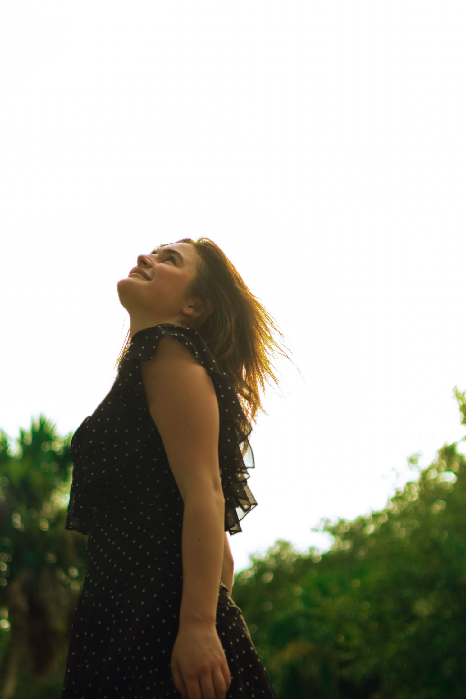 Young woman in black polka dot dress tilting head back outdoors with sunlit sky background.