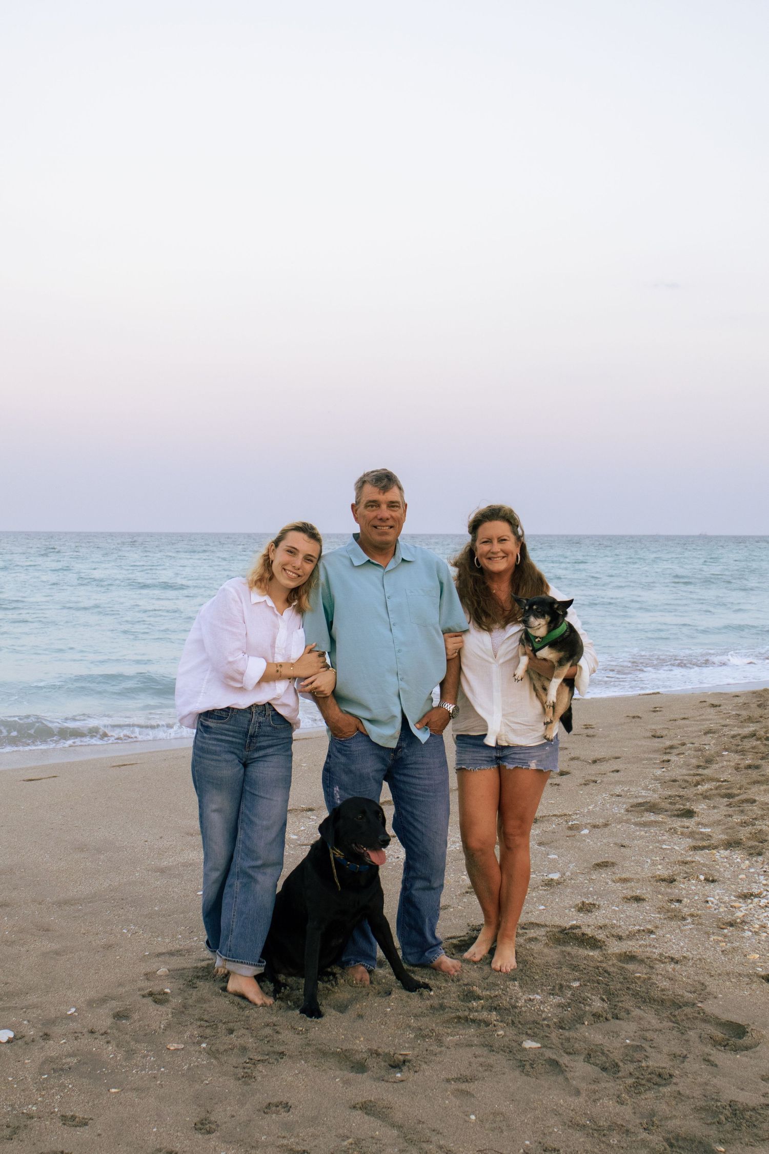 Family of three posing on beach at dusk with two dogs, ocean waves visible in background.