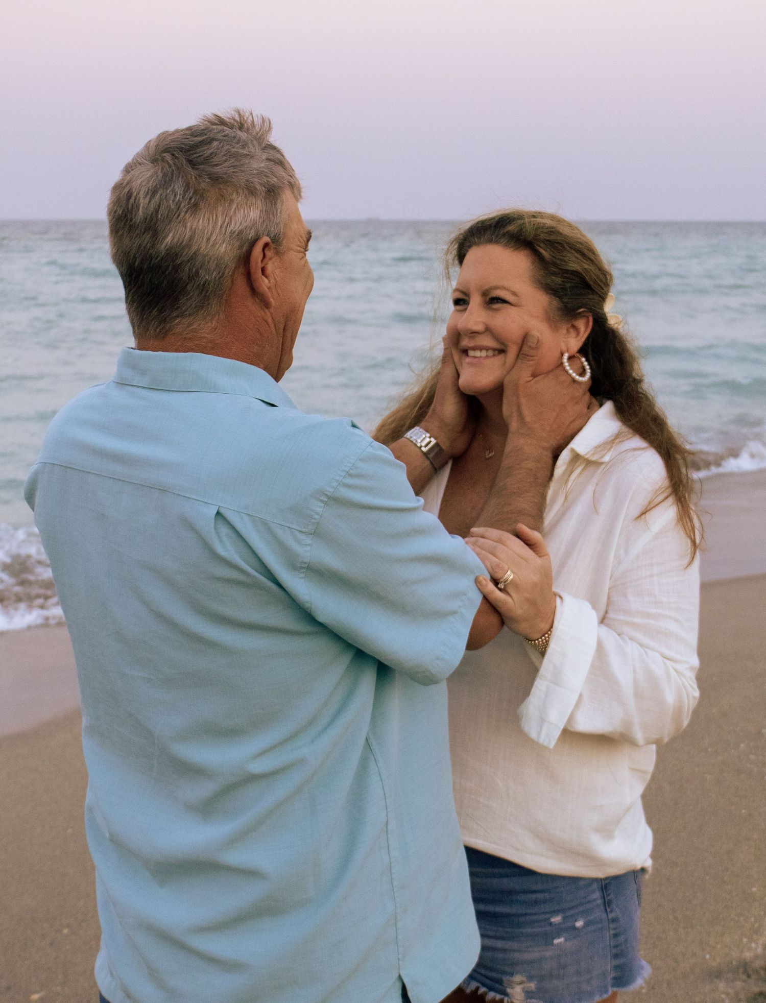 Couple sharing a romantic moment on the beach at dusk with ocean waves behind them.