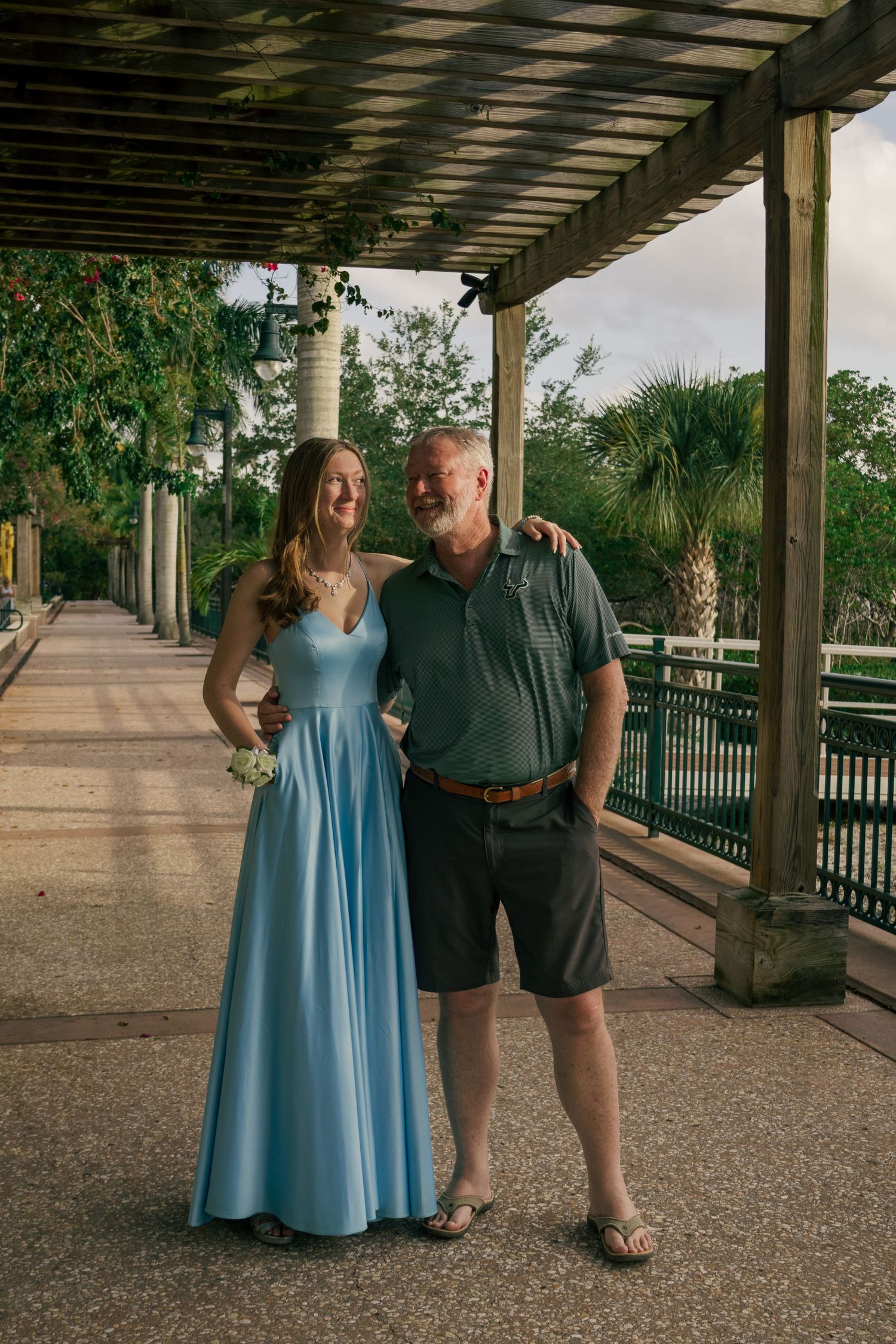 Young woman in light blue prom dress smiling with older bearded man under a pergola walkway.