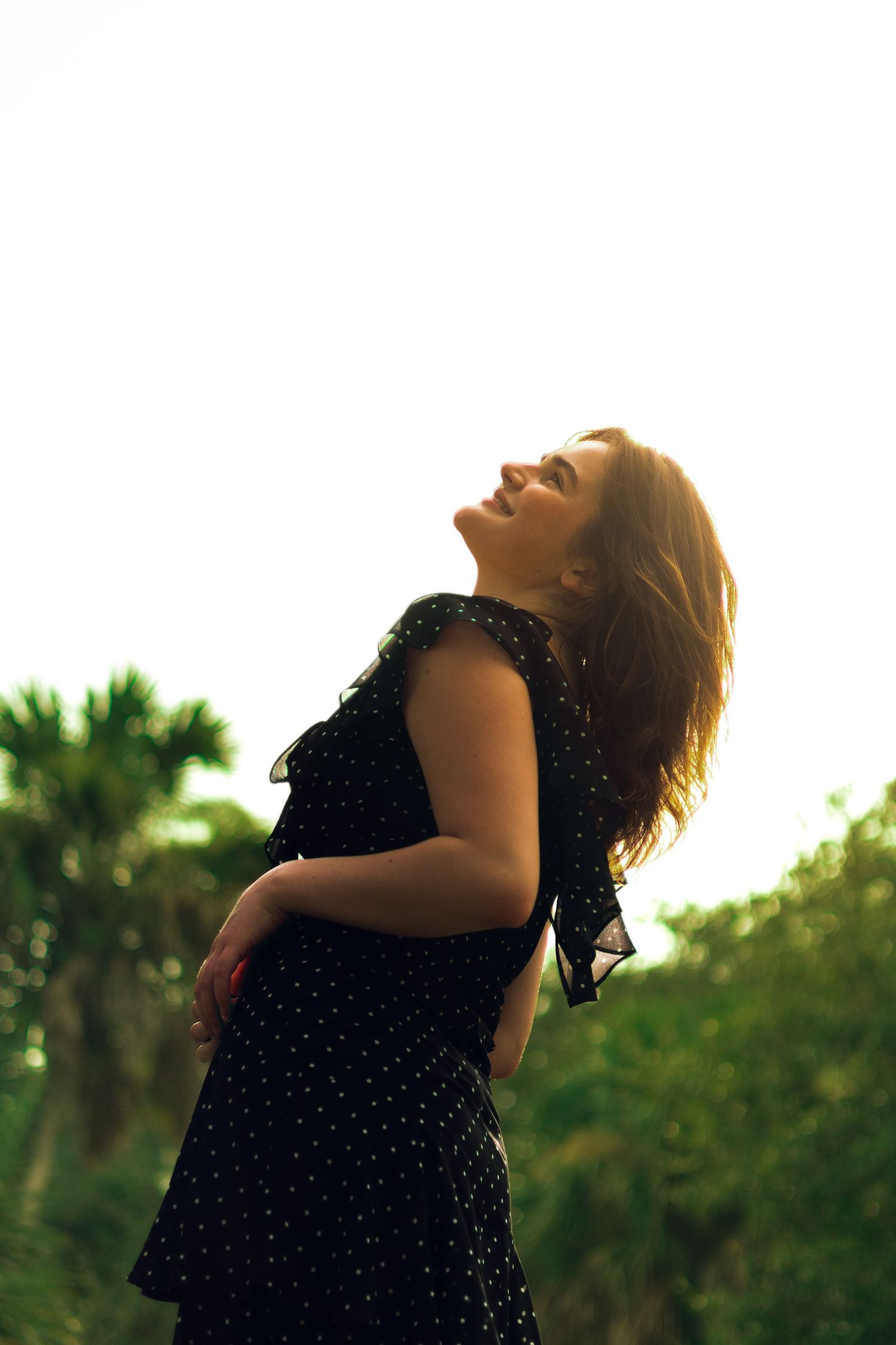 A young woman in a black polka dot dress stands outdoors, head tilted back, bathed in golden backlight.