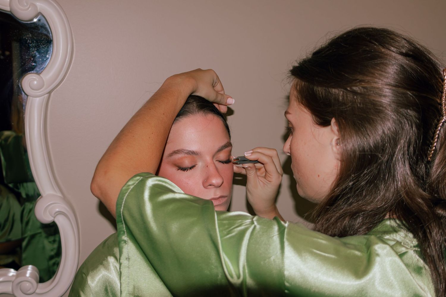 A makeup artist in a green satin robe applies eyeshadow to a woman in a matching robe before a wedding.