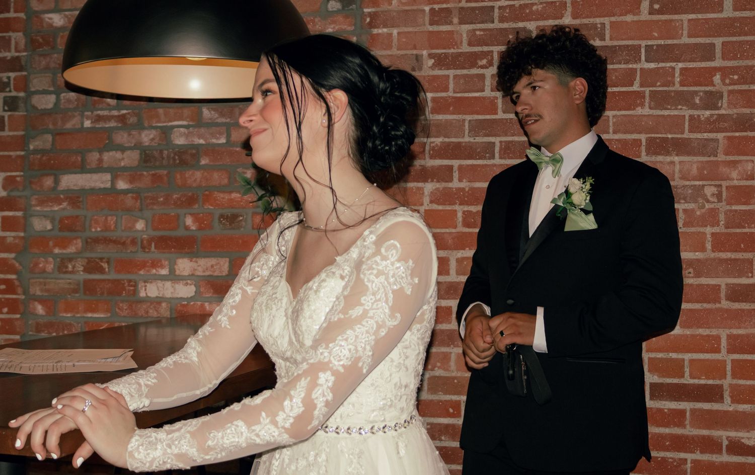 Bride in lace gown and groom in black tuxedo at rustic brick wall wedding venue.