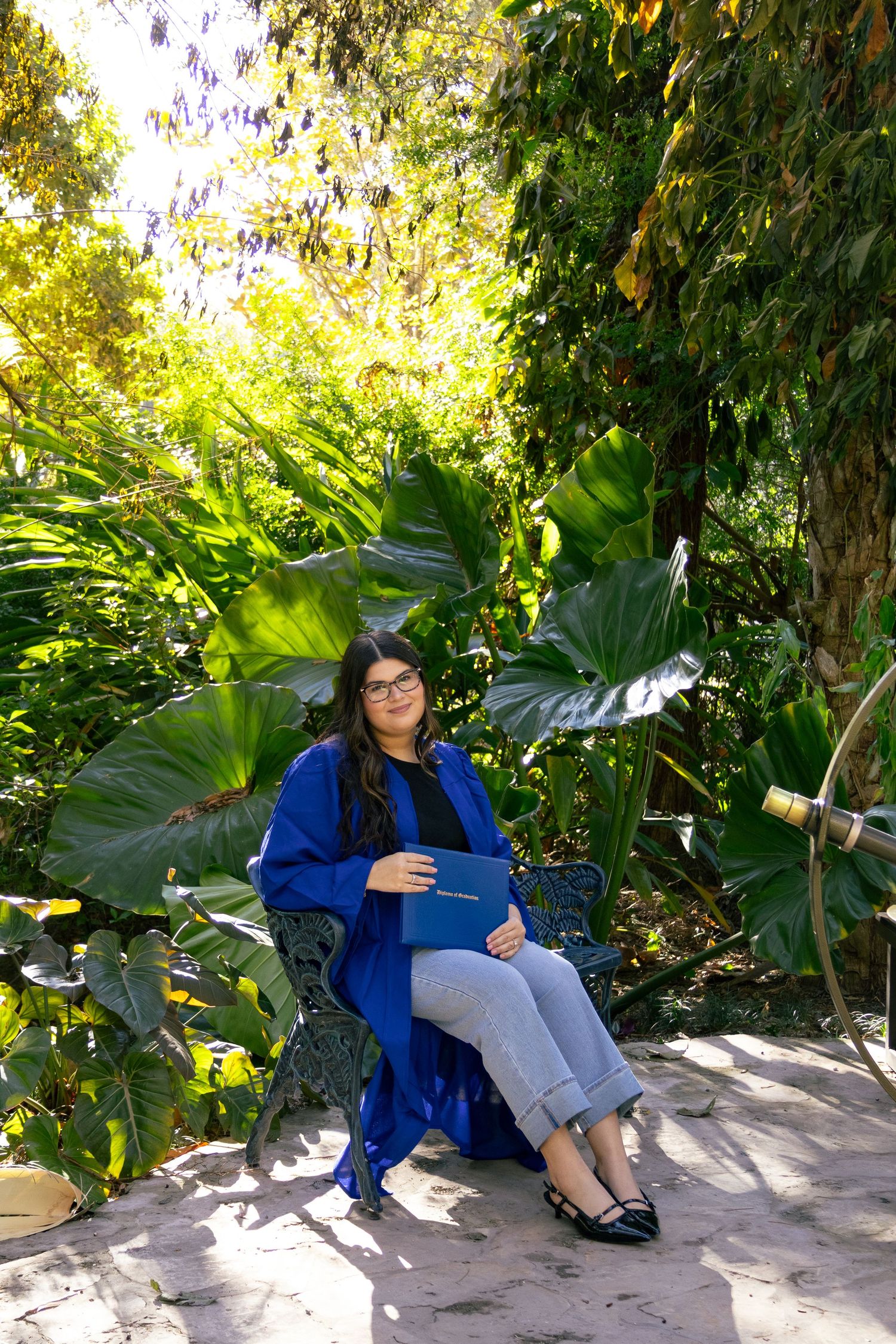 Graduate in blue gown holds diploma while seated on ornate bench in sunlit tropical garden.
