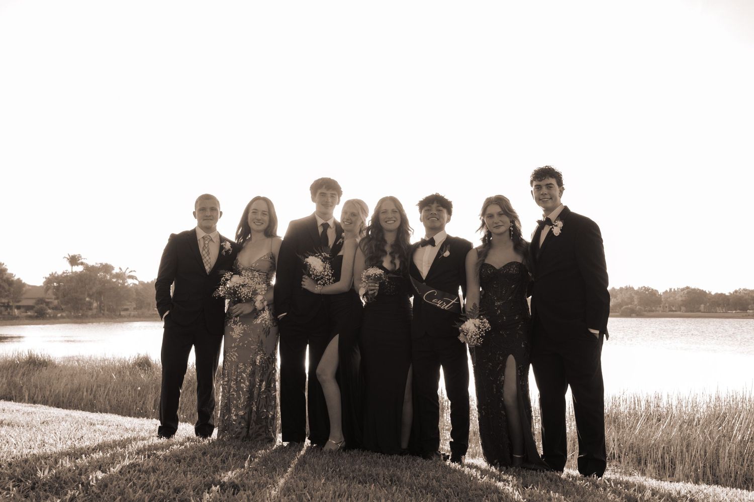 A sepia-toned photo of eight prom students posing together by a lake at golden hour.