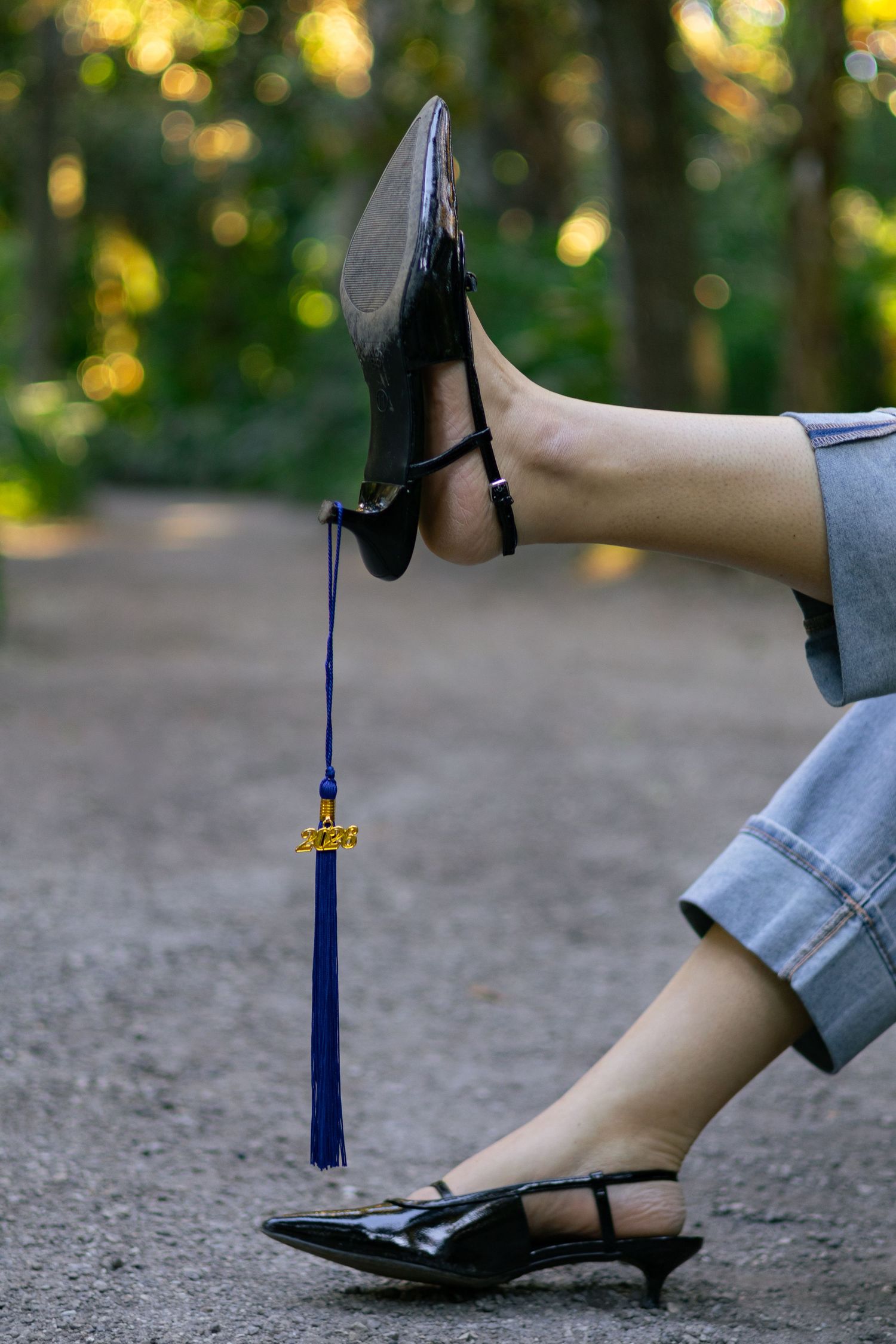 Person balancing a graduation cap on their foot with a blue tassel hanging down, seated outdoors on a gravel path.