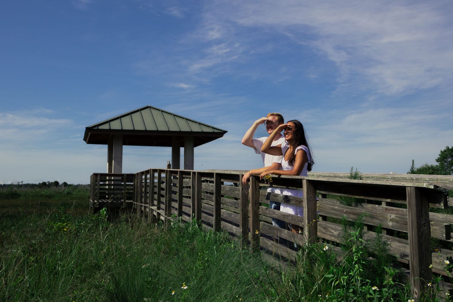 Couple sitting on wooden fence railing at outdoor boardwalk overlook under bright blue summer sky.